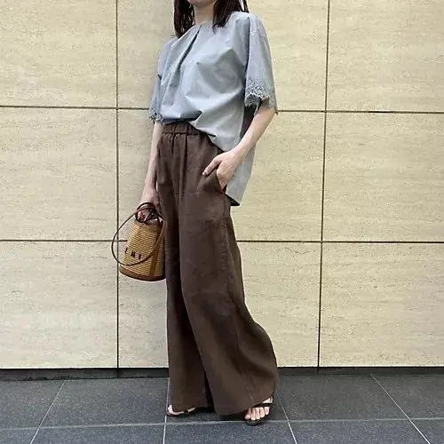 グレーのゆったりしたワンピースを着た女性が、木製の床の上に立ち、白いトートバッグを左手に持ち、笑顔でカメラを見つめている