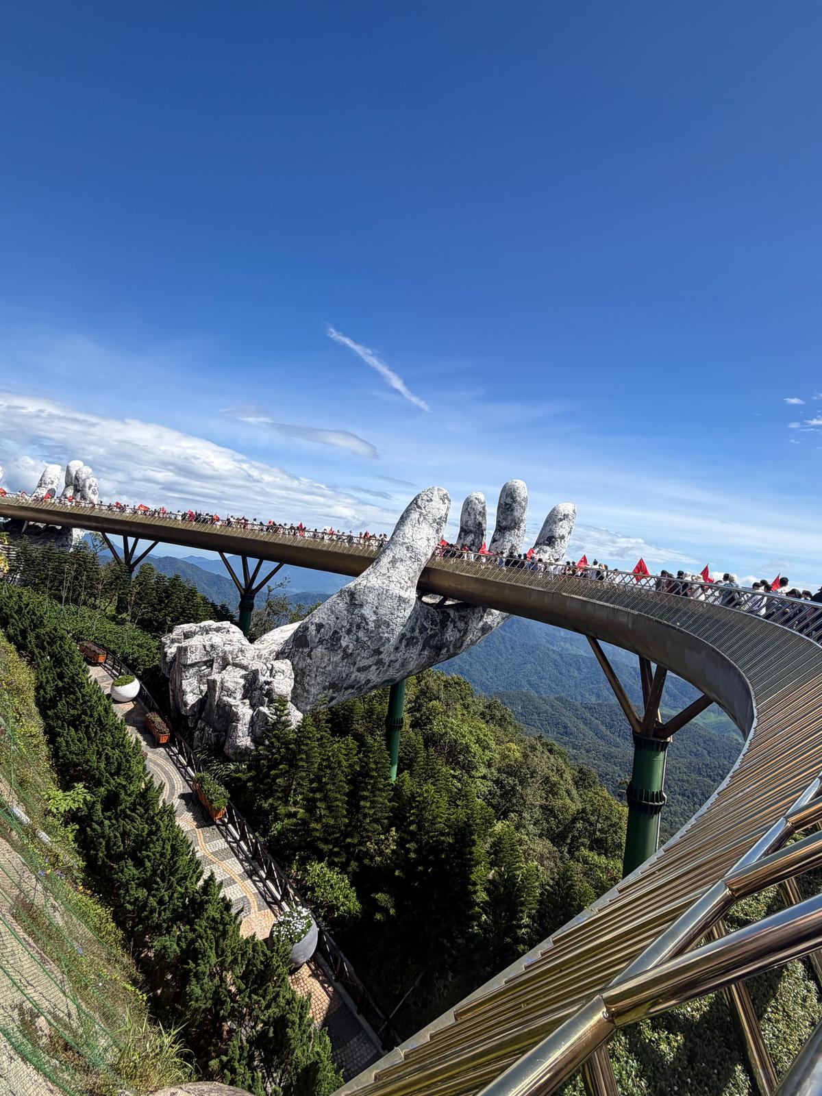 ベトナム・ダナンの人気観光地「神の手橋」。巨大な石の手が空中にそびえ立ち、指先から橋が伸びるユニークな景観が広がる。緑豊かな山々を背景に、赤い帽子をかぶった人々が橋を歩いている。青空に白い雲が浮かぶ。
