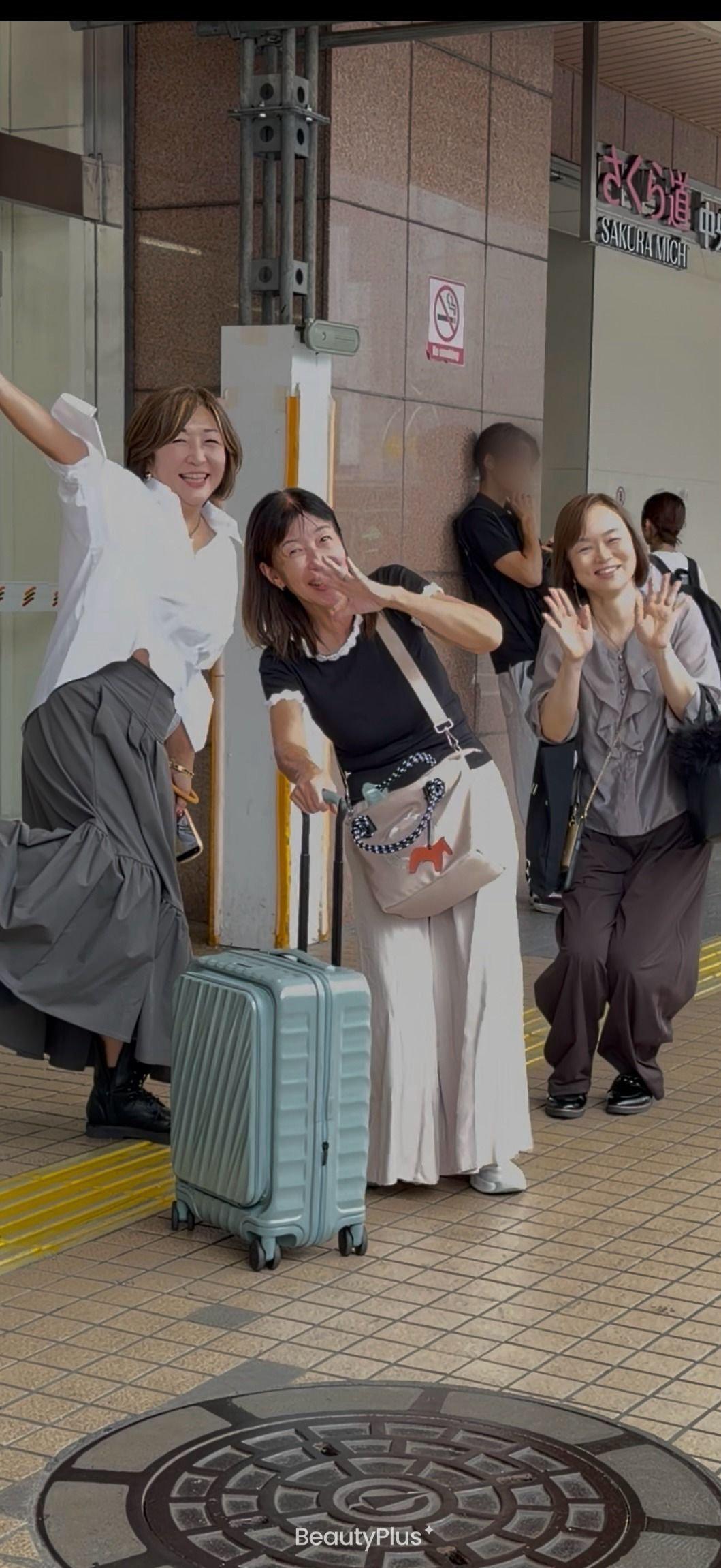 3人の女性が笑顔で手を振っている写真。左から2人目の女性は黒いトップスと白いスカート、右隣の女性は白いロングスカートにベージュのバッグ、左端の女性はグレーのスカートを着用。背景には建物の壁と「SAKURA MICH」の看板が見える。