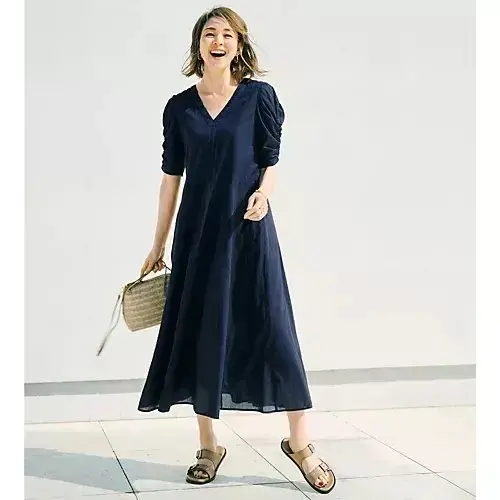 ダークブルーのVネックロングドレスを着た女性が、籐のトートバッグを持ち、笑顔で歩いている。足元はサンダル。