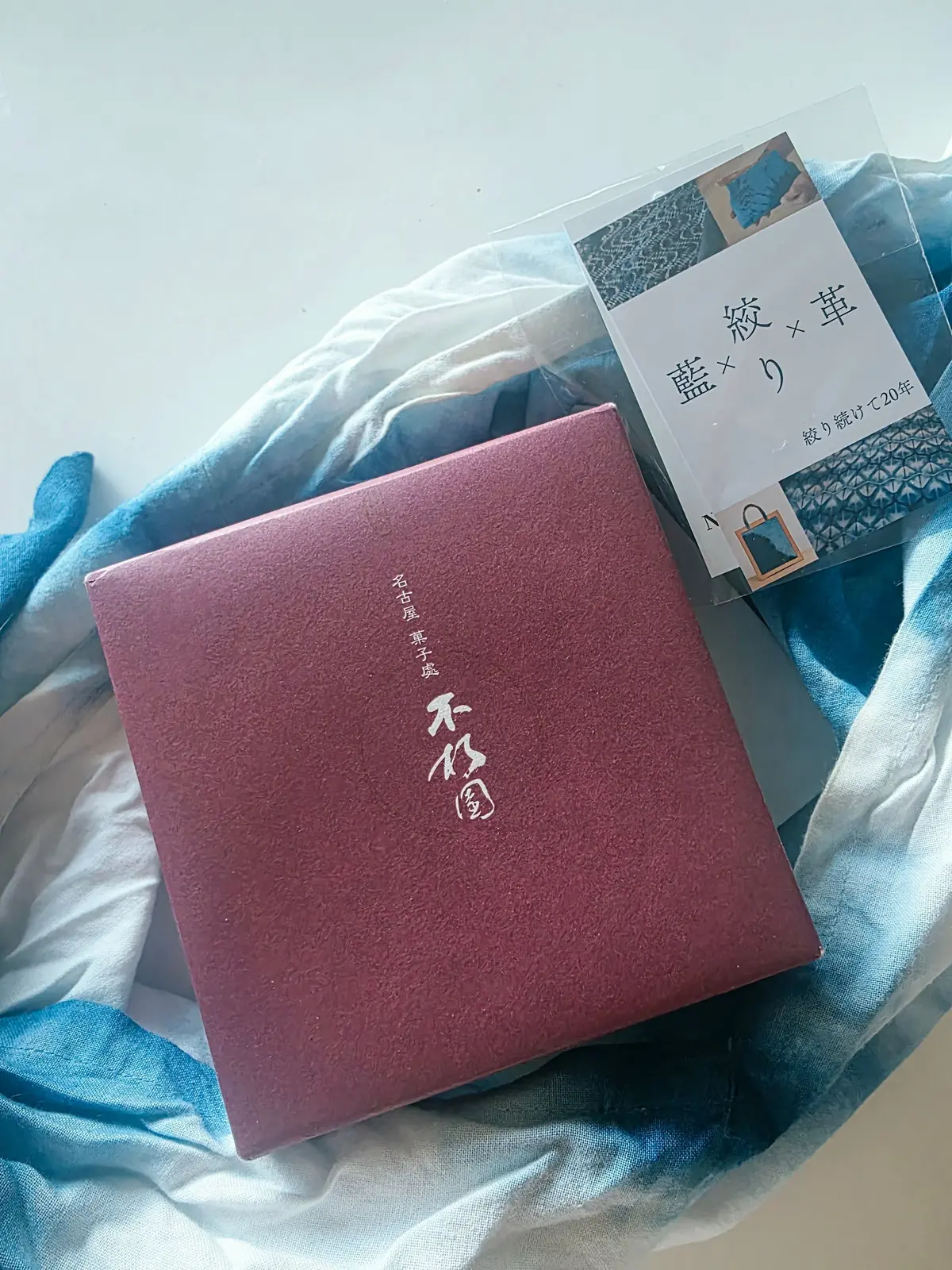 藍染め製品のボックスと布が置かれた写真。左には濃い赤色の箱、「不朽園」の文字が白で表記されている。右には藍色の箱で、「藍×藍」と書かれた説明書が添えられている。背景には、様々な色合いの藍染めの布が散らばっている。