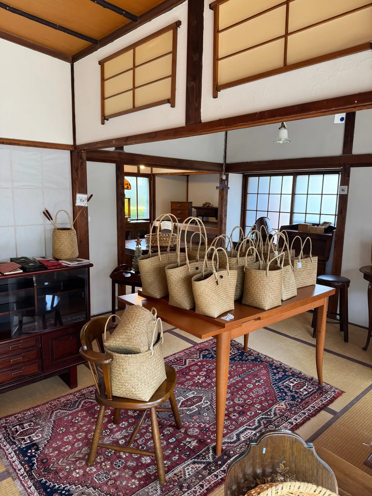 木造の梁がある広々とした空間に、複数の麦わら製バッグがテーブルに並べられている。背景には障子や木製の家具が見え、伝統的な日本の家屋の様式である。テーブルには赤い本が置かれ、手すり付きの椅子が配置されている。