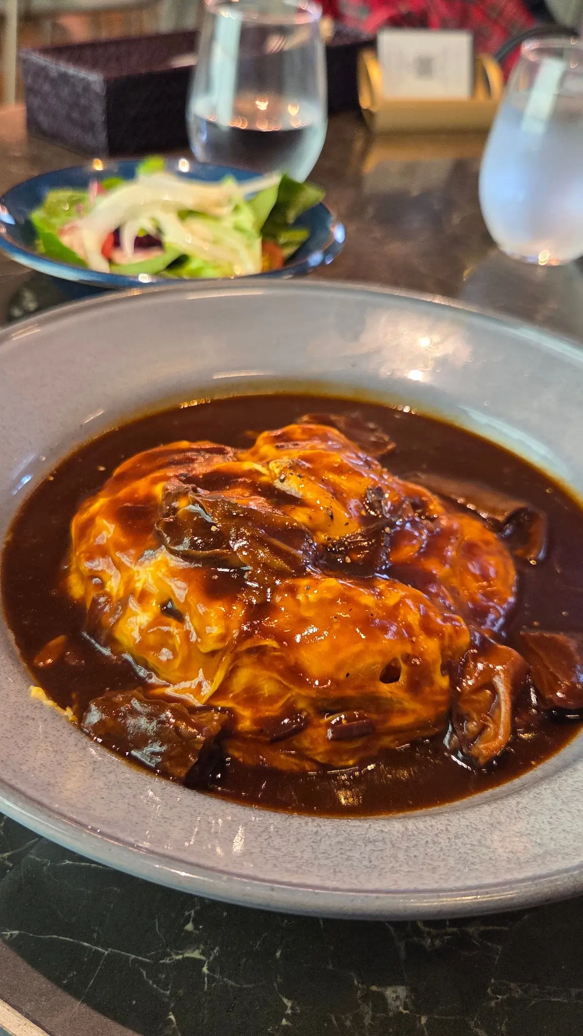 鶏肉の煮込みが茶色いソースで覆われた皿、横に青野菜のサラダとガラスの飲み物が置かれたテーブル