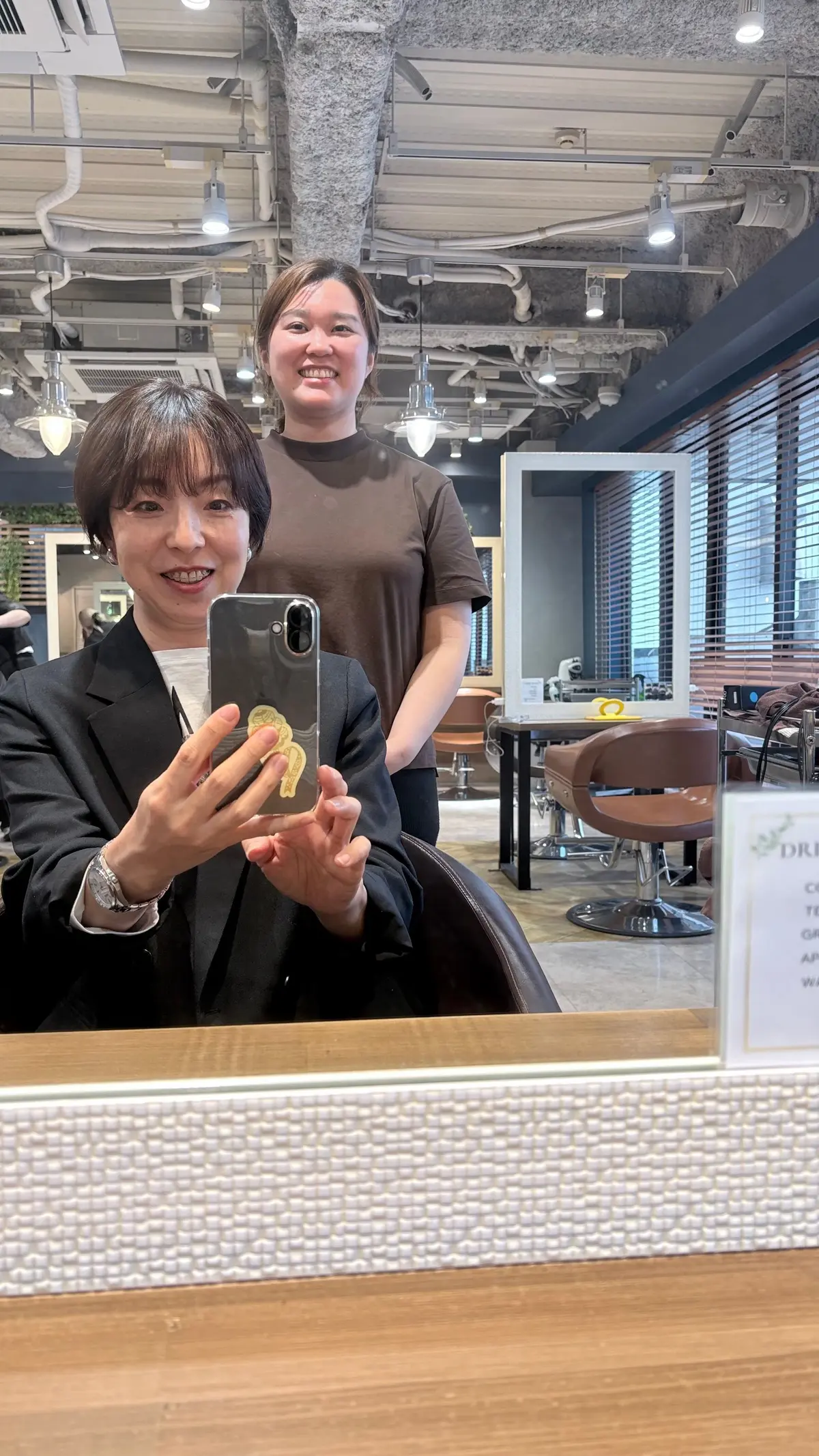 美容室のインテリアと鏡のあるカウンターで、2人の女性が映る。前景の女性は黒のスーツで携帯電話を操作し、背景の女性は茶色のTシャツを着ている。