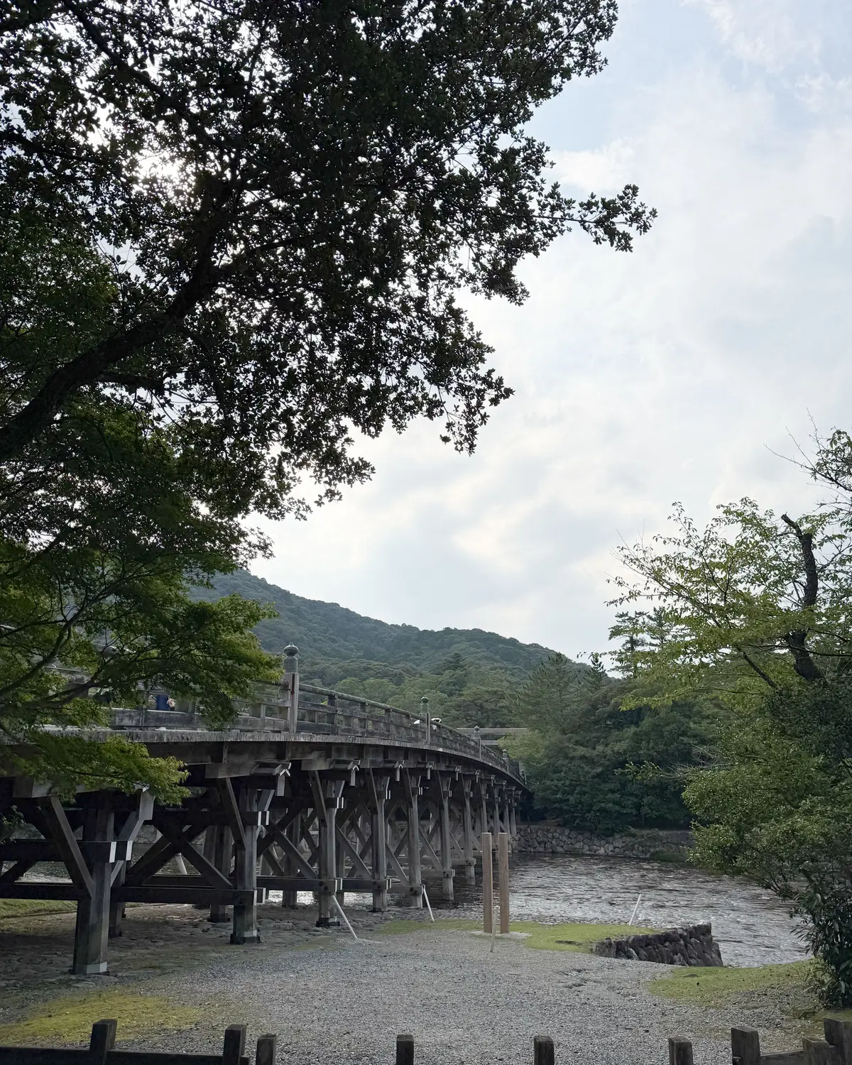 木造の橋が川を渡り、背景には緑豊かな山々が広がる風景。橋脚は木組みでできており、川面は流れが速い。手前に木製の柵がある。