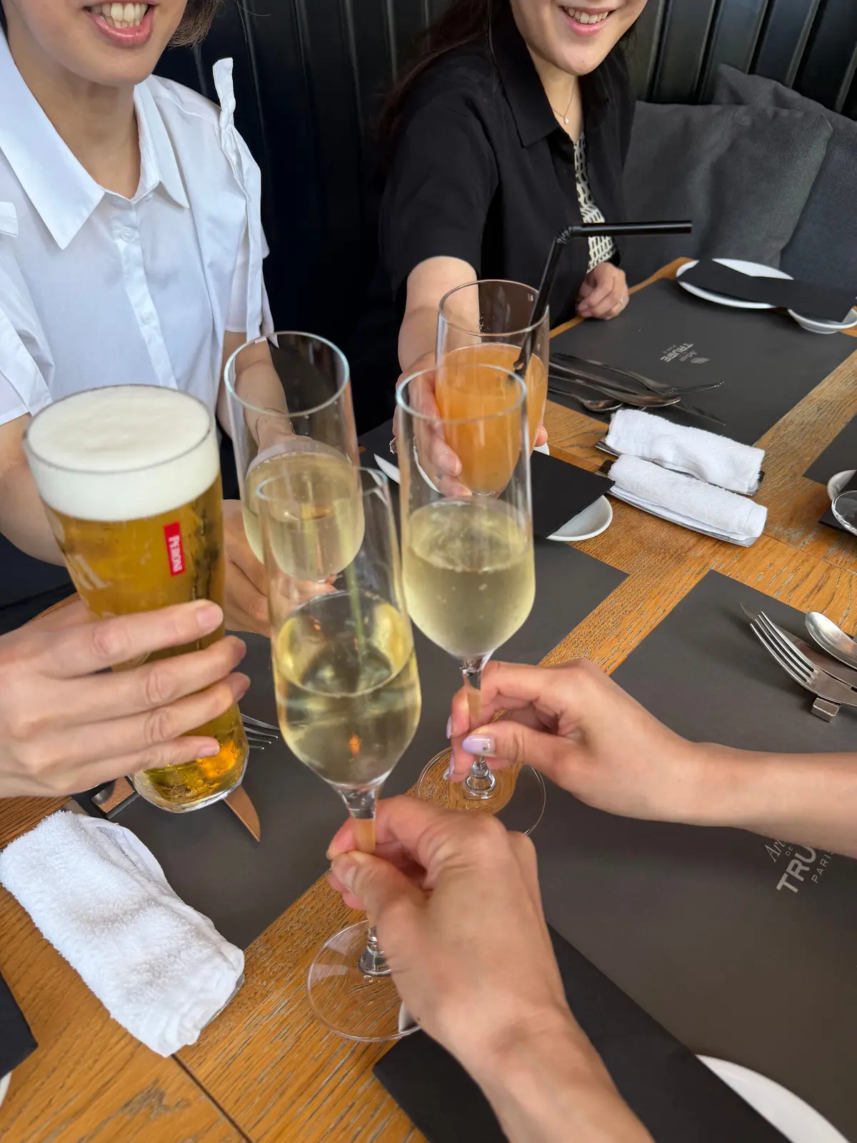 テーブル席で、複数の人がビールやスパークリングワインをグラスに注ぎ、乾杯を楽しんでいる様子。木製のテーブルに置かれた黒いプレースマットと、白いナプキンが目立つ。