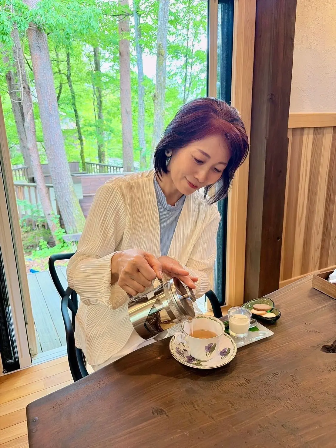 女性が木製のテーブルに座り、花模様のティーカップに茶を注いでいる。窓の外には緑豊かな木々が広がる。