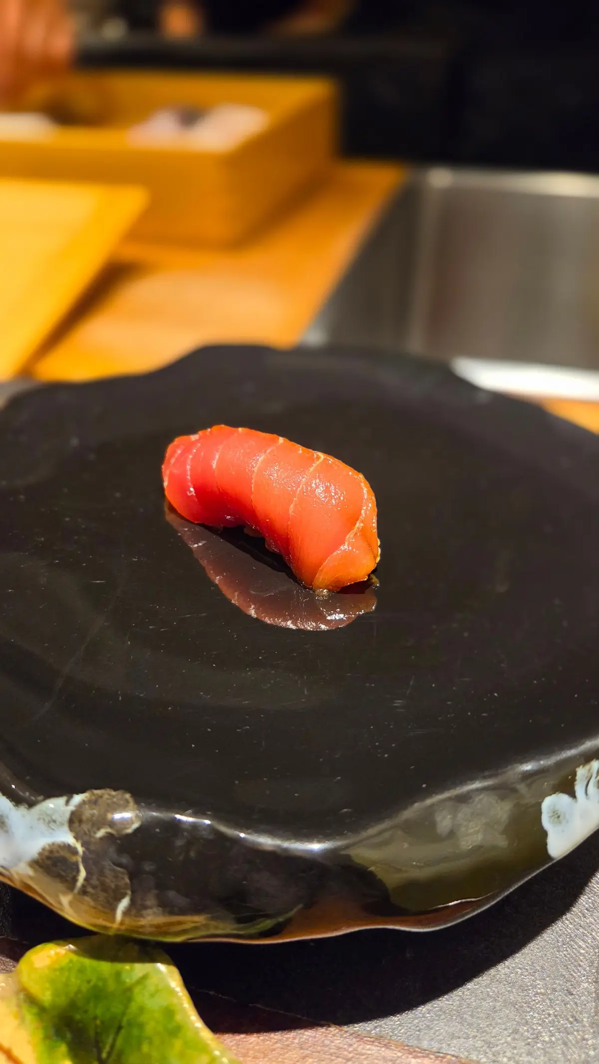 黒いお皿に盛り付けられた、赤身のマグロ寿司。表面は照りがあり、断面が渦巻き状になっている。お皿の脇には緑色の薬味。