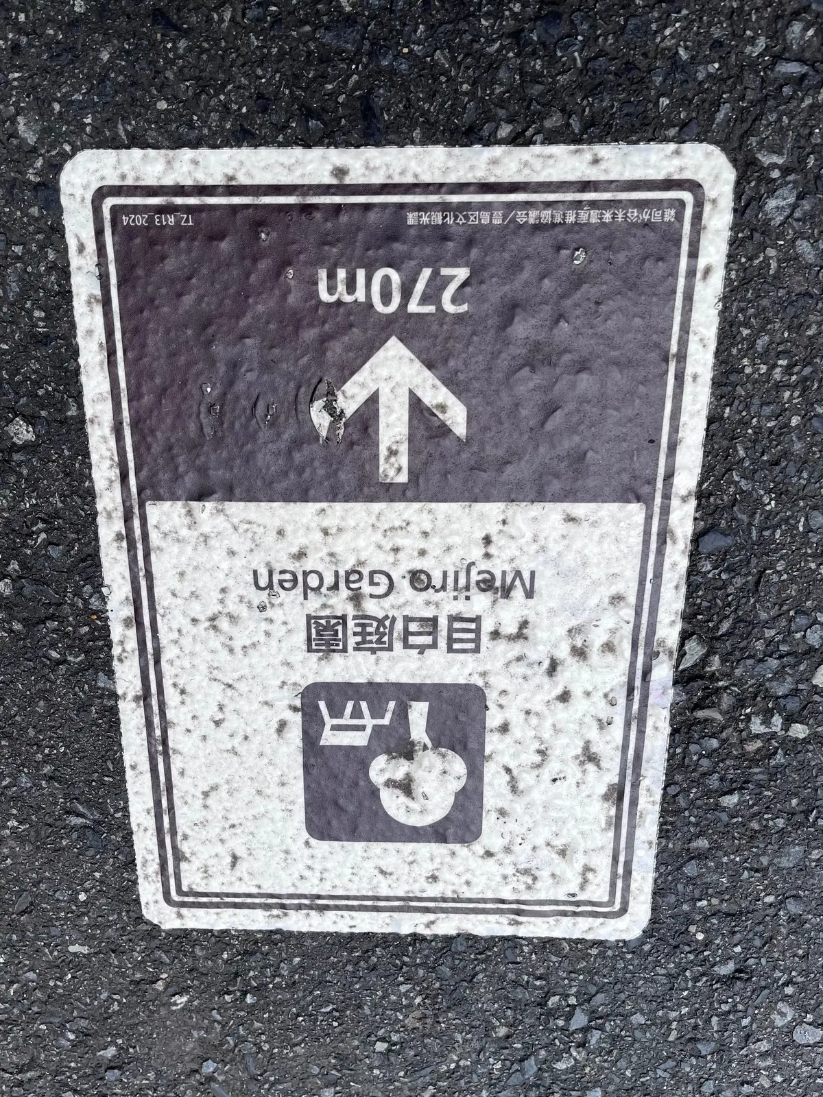 灰色の屋根と白い壁を持つ伝統的な日本建築の案内所。正面には石畳の広場があり、両脇に案内板が置かれている。背景には緑豊かな木々が茂り、青空が広がっている。