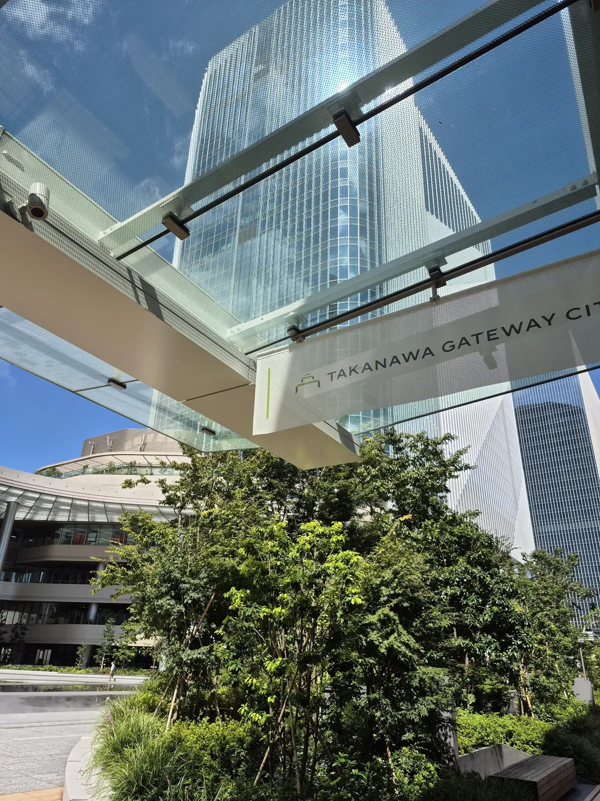高層ビル群と緑豊かな植栽が調和した都市景観。Takanawa Gateway Cityの案内板が設置された歩道の上から、ビルの窓面が青空に映り込んでいる。