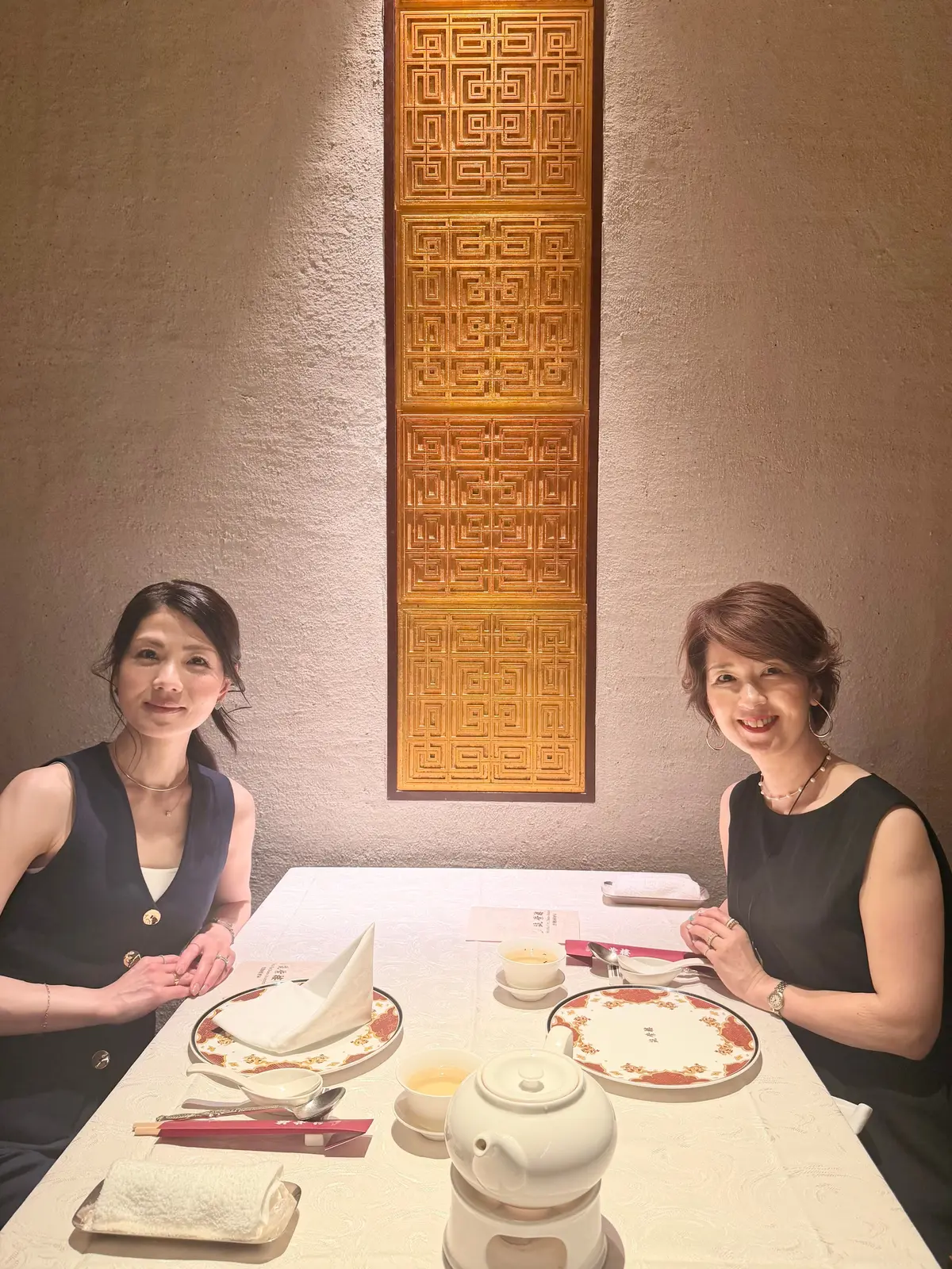 二人の女性がテーブルに座り、茶器や料理が配置されたテーブルで会話する様子。背景には幾何学模様の木製パネルが飾られている。