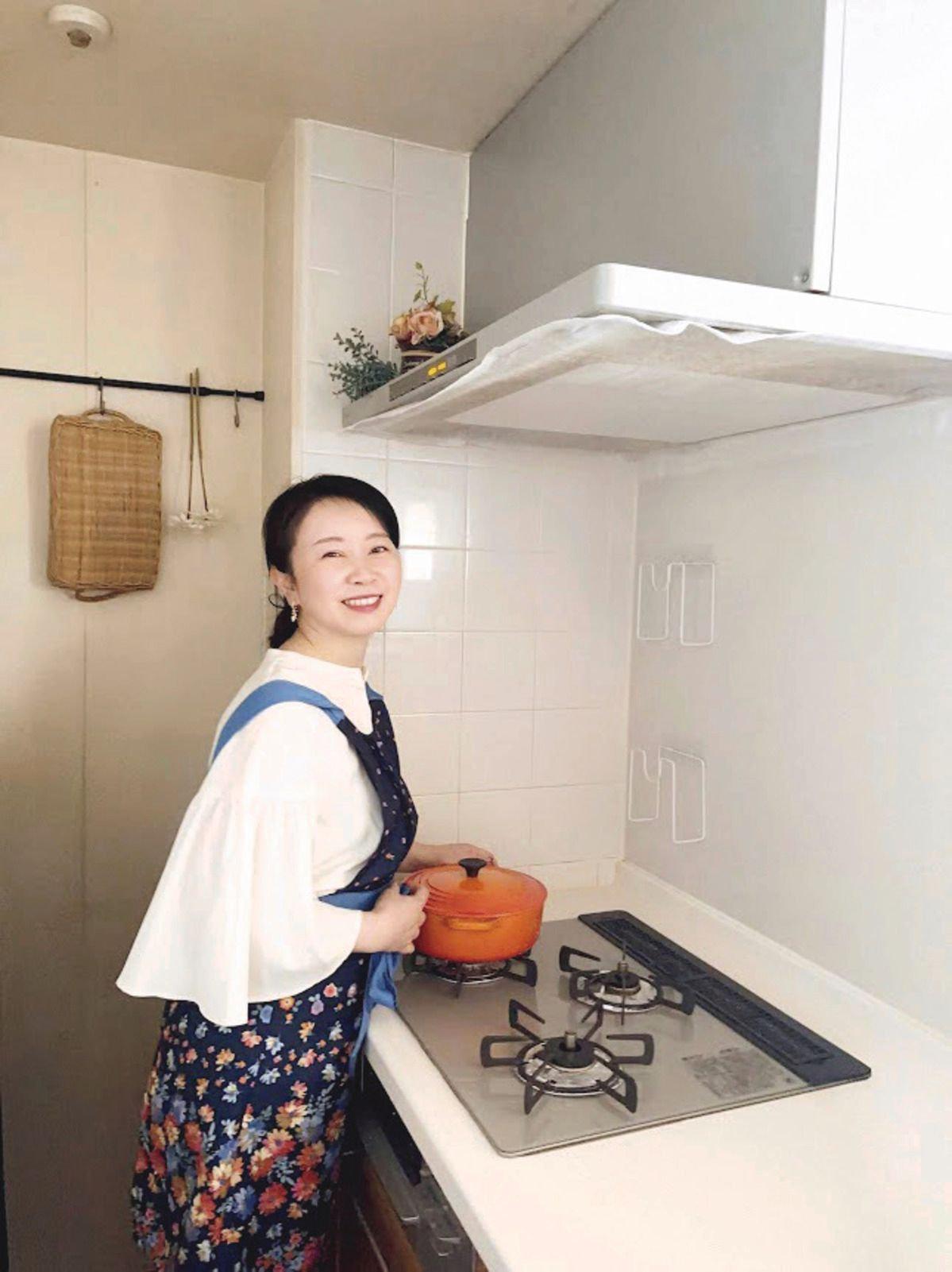 笑顔の女性が、白いトップスと花柄のスカートを身につけ、キッチンで鍋を持ちながら立っている。背景には白いタイルと換気扇、調理台がある。