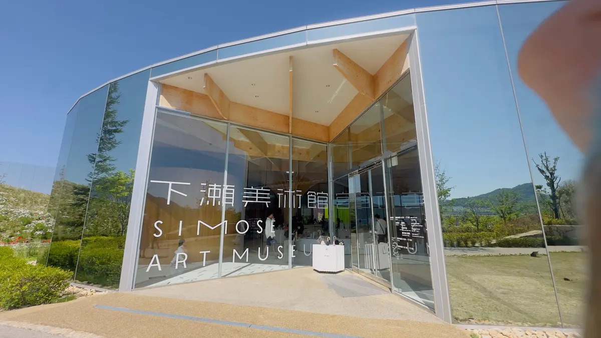 下浦美術館のガラス張りの建物で、木製の天井が内観から見える。緑の芝生と木々が背景にある。