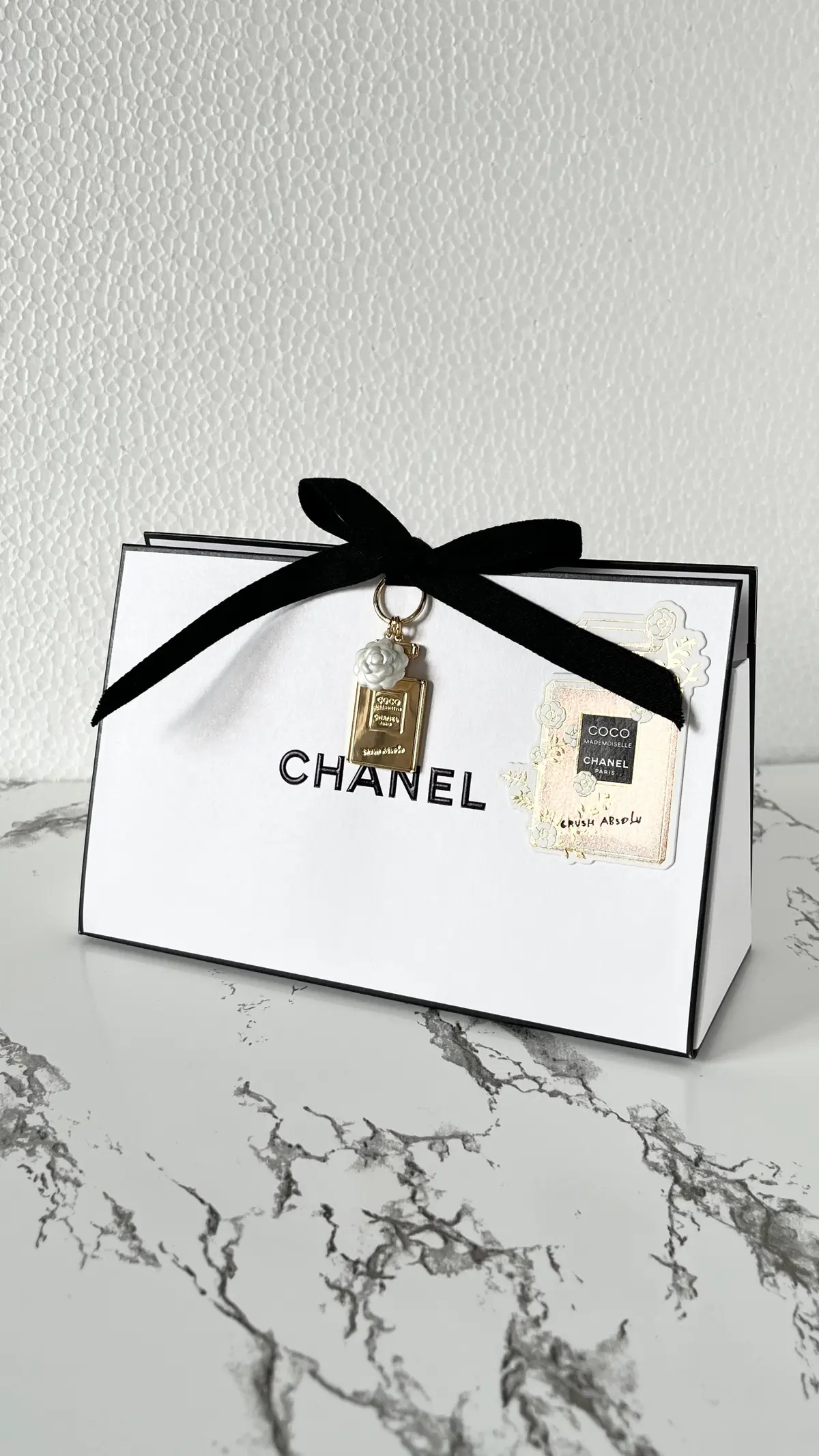 白い箱に「CHANEL」のロゴがプリントされ、黒いリボンで飾られています。箱には、シャネルの香水やコスメのキーホルダーが2つ、リボンを通して吊り下げられています。背景は白い壁と大理石調のテーブルです。