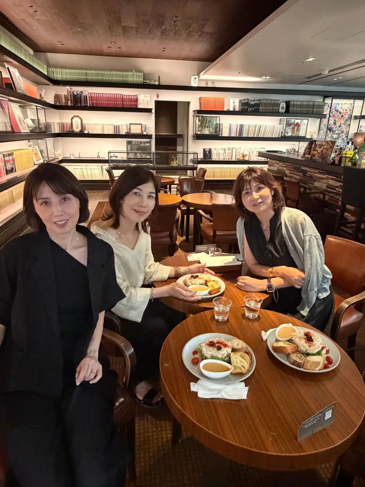 カフェのような空間で、3人の女性が丸テーブルを囲んで食事を楽しんでいる様子。左から黒いジャケット、白いシャツ、グレーのニットを着ており、それぞれ笑顔を見せている。テーブルには前菜の盛り合わせと飲み物が置かれている。背景には本棚や装飾が配置されている。