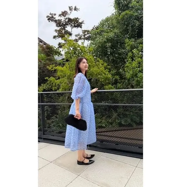 青いドット柄のワンピースを着た女性が、手すりに寄りかかりながらこちらを見ている。黒いパンプスを履き、黒いクラッチバッグを持っている。背景には緑豊かな植物が見える。
