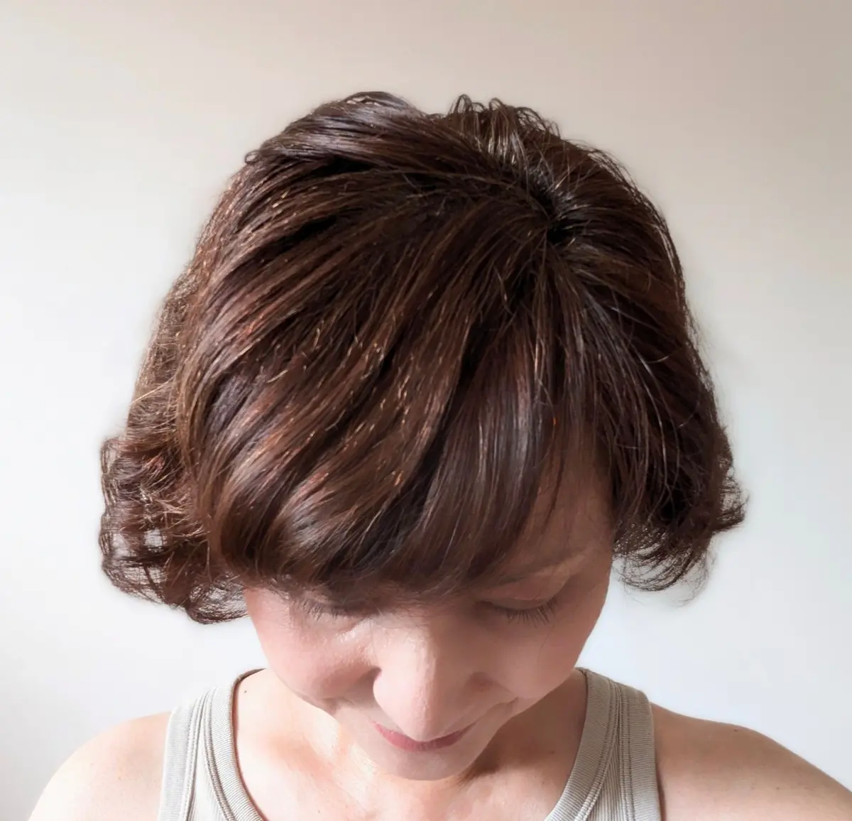 茶色のボブヘアに前髪をつけた女性が、白い壁の前で俯いている。グレーのタンクトップを着ており、顔は影になっている。