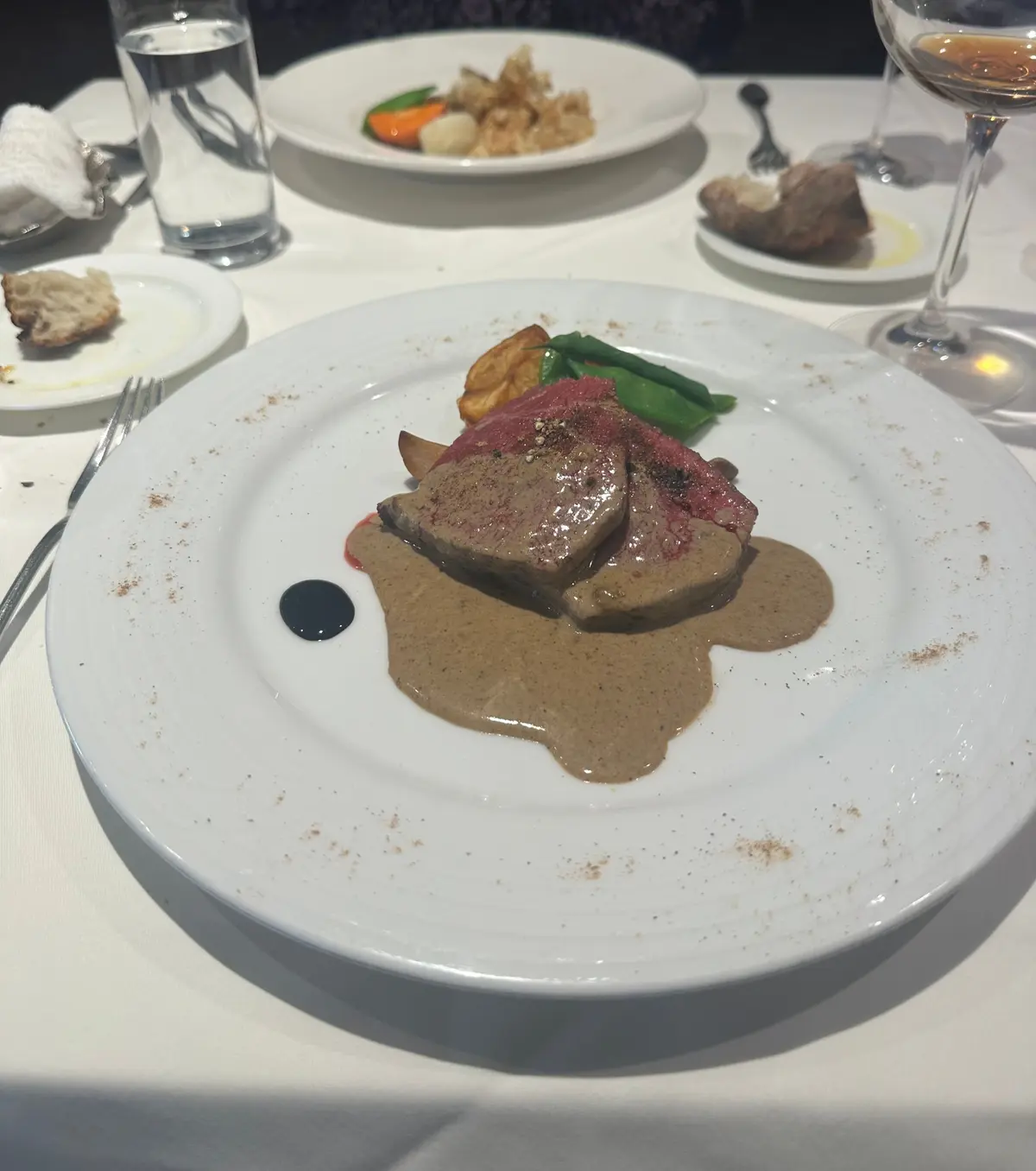 白テーブルに置かれた、肉料理の盛り合わせ。肉は薄切りで、ソースをかけてあり、付け合わせにポテトと野菜、赤ワイングラスが写っている。