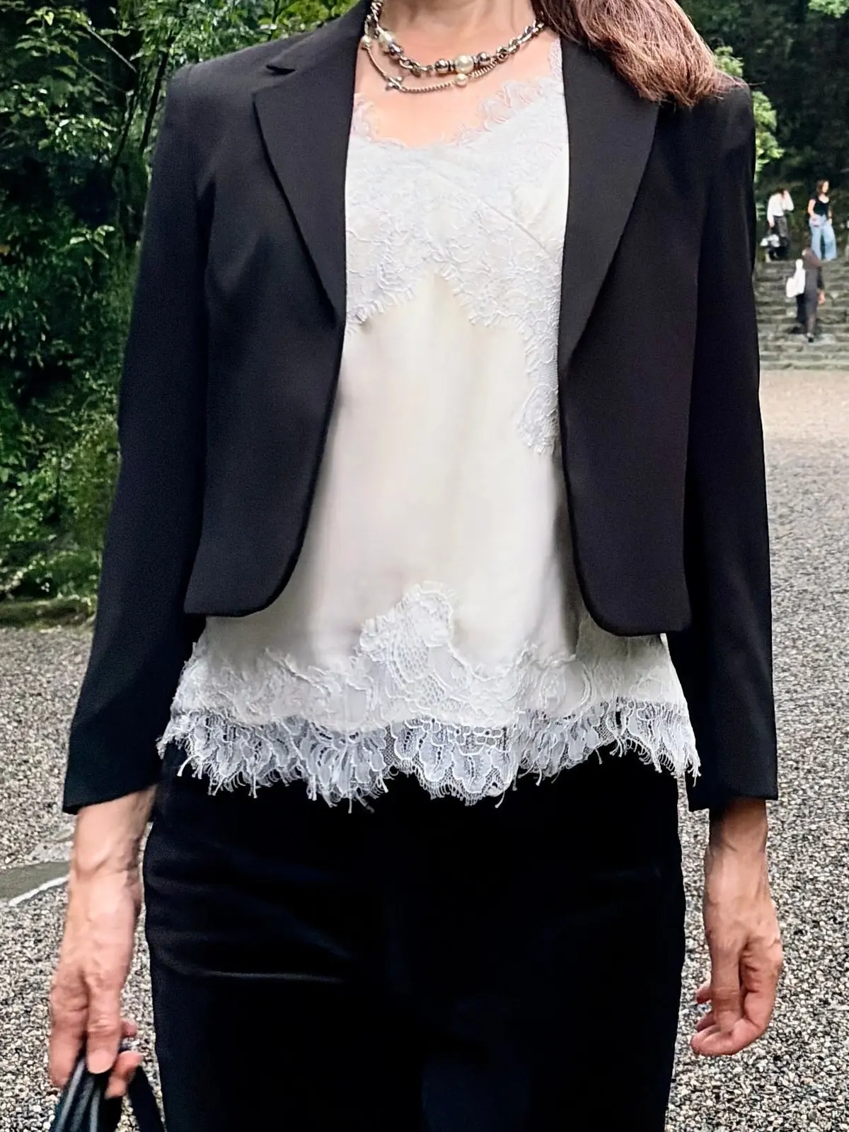 黒のジャケット、白いレースのトップス、黒いパンツを着用した女性。首にはパールとビーズのネックレス。背景には緑の木々と石畳の道。