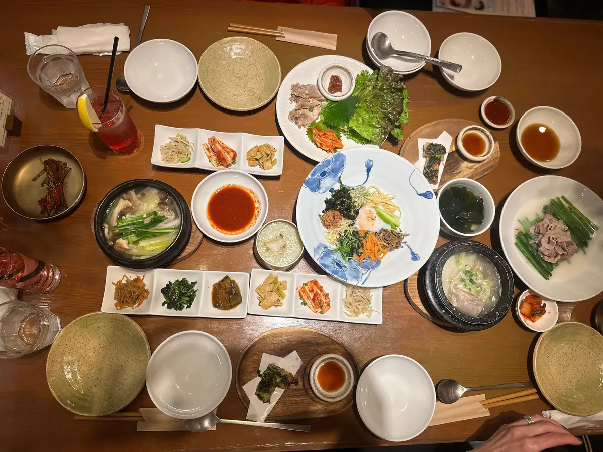 テーブルに並んだ韓国料理の盛り合わせ。様々な小皿料理（キムチ、おかず）、スープ、飲み物、食器が配置されている。中央には、韓国風サラダの大きな皿がある。