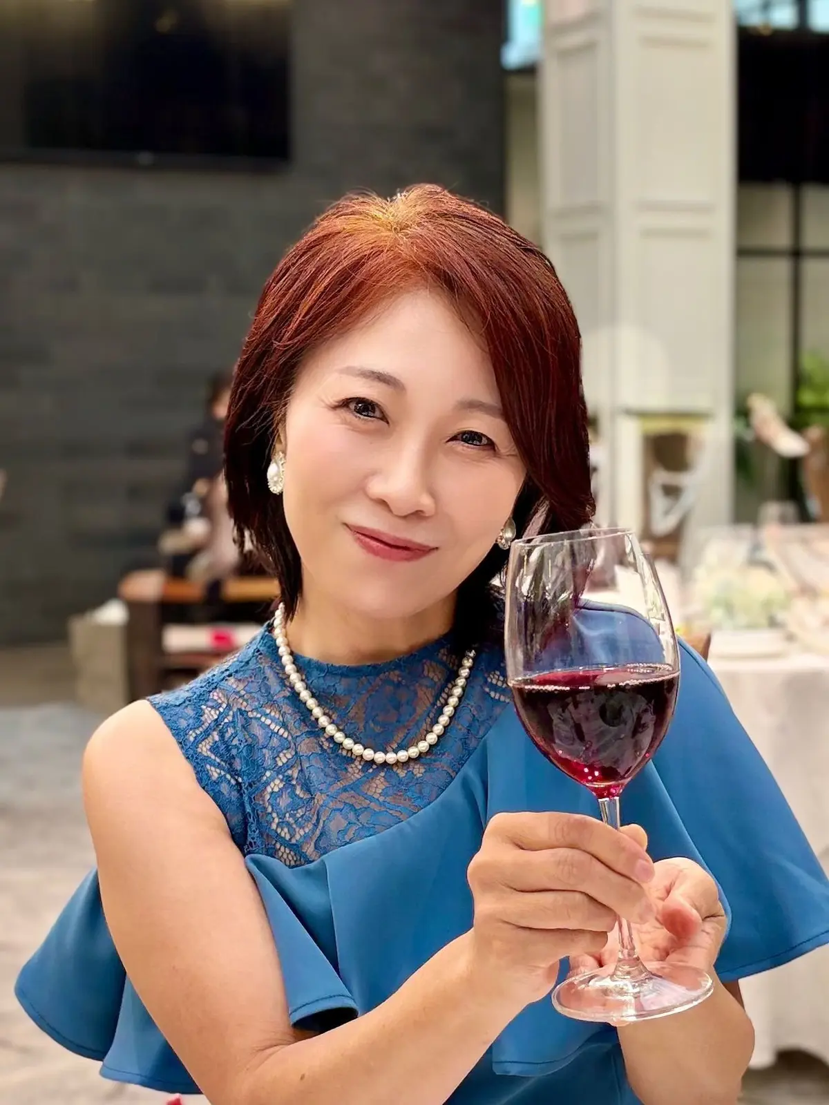 赤毛の女性がブルーのレースドレスと真珠のネックレスを着用し、赤ワインをグラスに注いでいます。