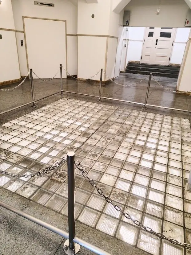 広々とした廊下の床に設置された、ガラスブロックでできた正方形のエリア。周囲には金属製のチェーンと支柱が置かれ、立ち入り禁止にされている。奥には白いドアと階段がある。