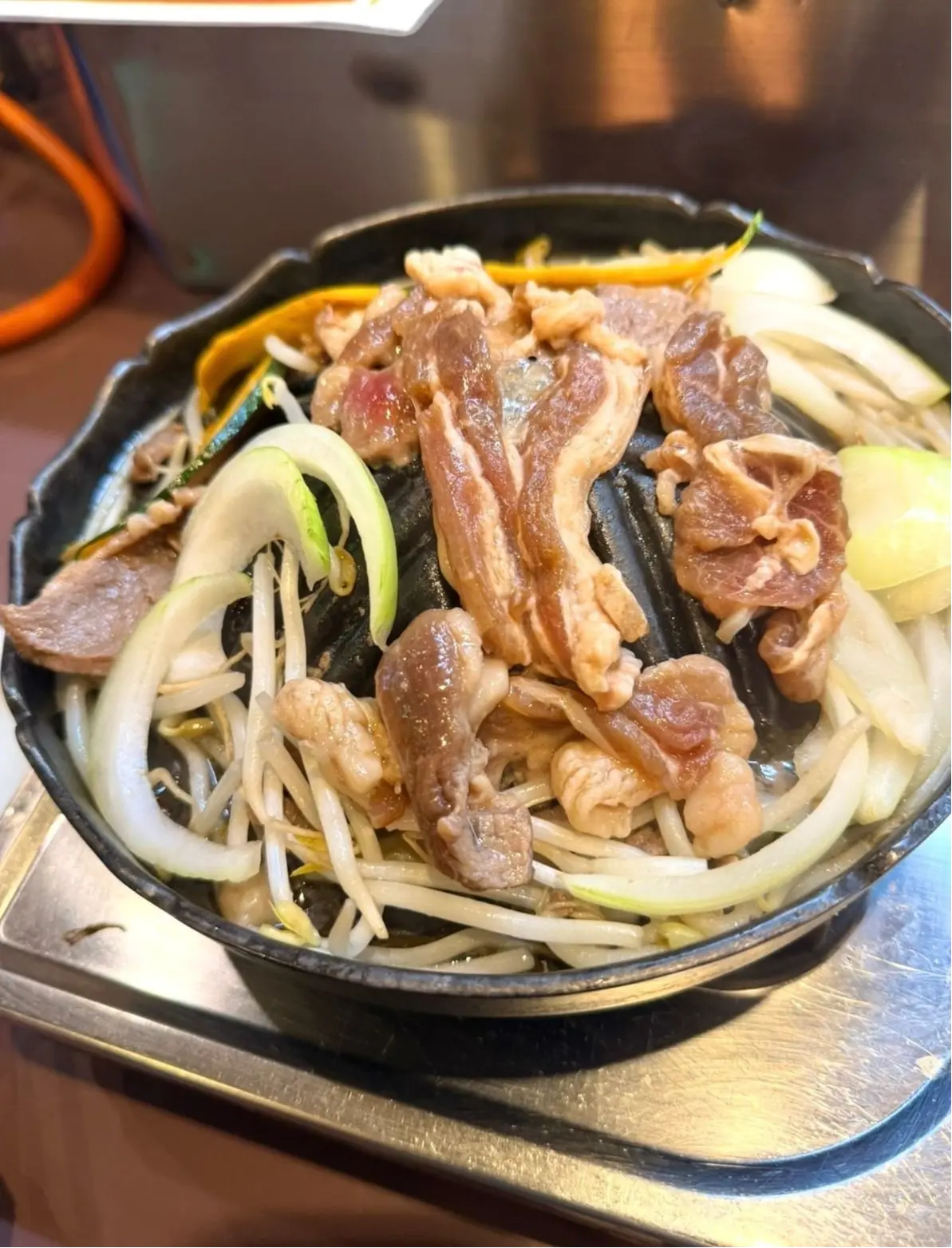 黒い鉄製のプレートに盛り付けられた、薄切り肉と野菜の炒め物。肉は淡いピンク色で、もやしや玉ねぎ、ネギなどの野菜が添えられている。