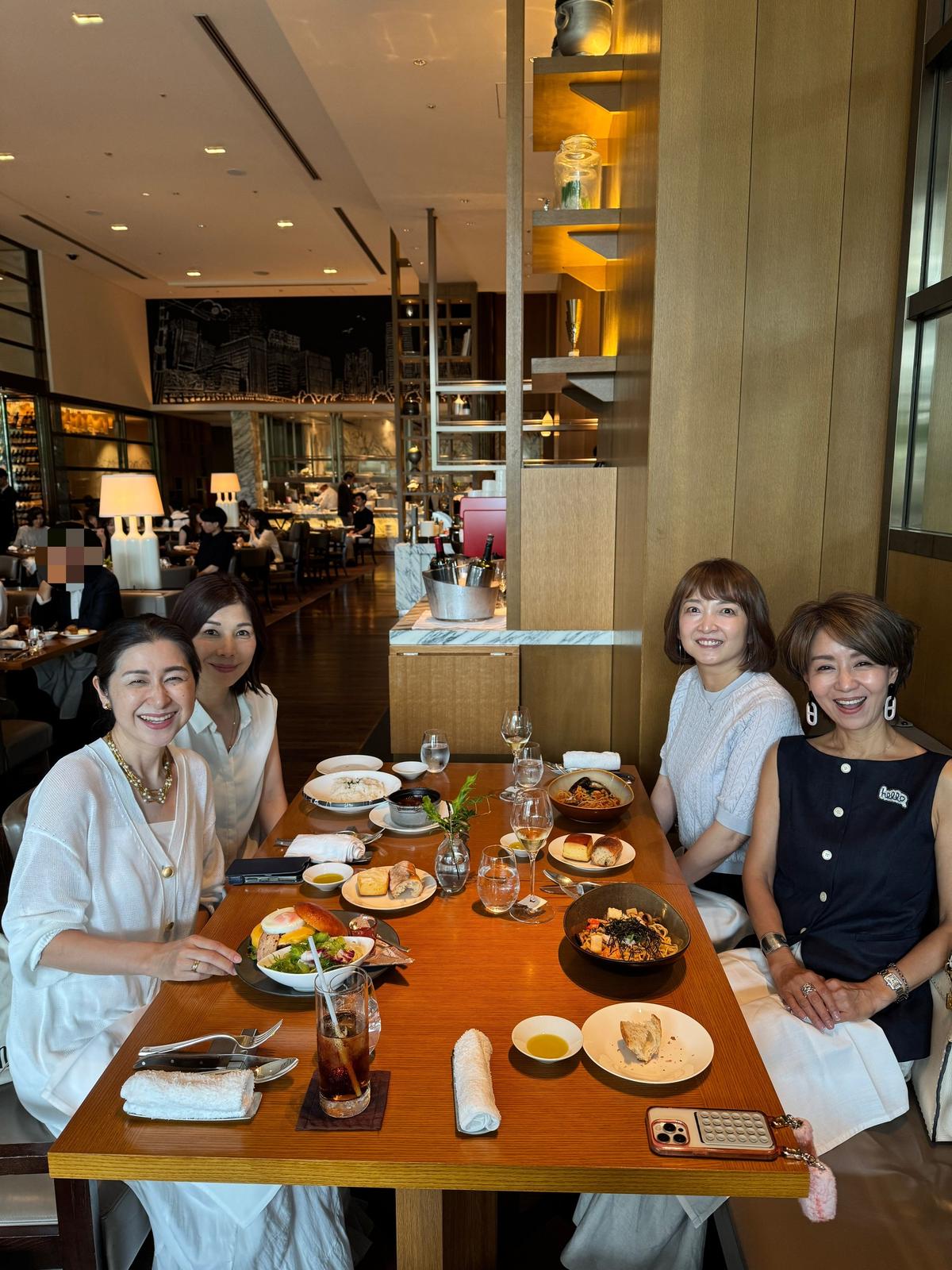 レストランで食事を楽しむ4人の女性。木製のテーブルには様々な料理とグラスが並び、背景には都市の壁画が見える。笑顔の女性たちが楽しげな雰囲気を醸し出している。