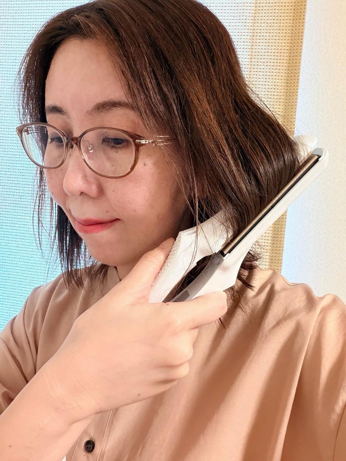 茶色の髪に眼鏡をかけた女性が、白いヘアアイロンで髪を整えている。ベージュのシャツを着ており、背景にはカーテンが見える。
