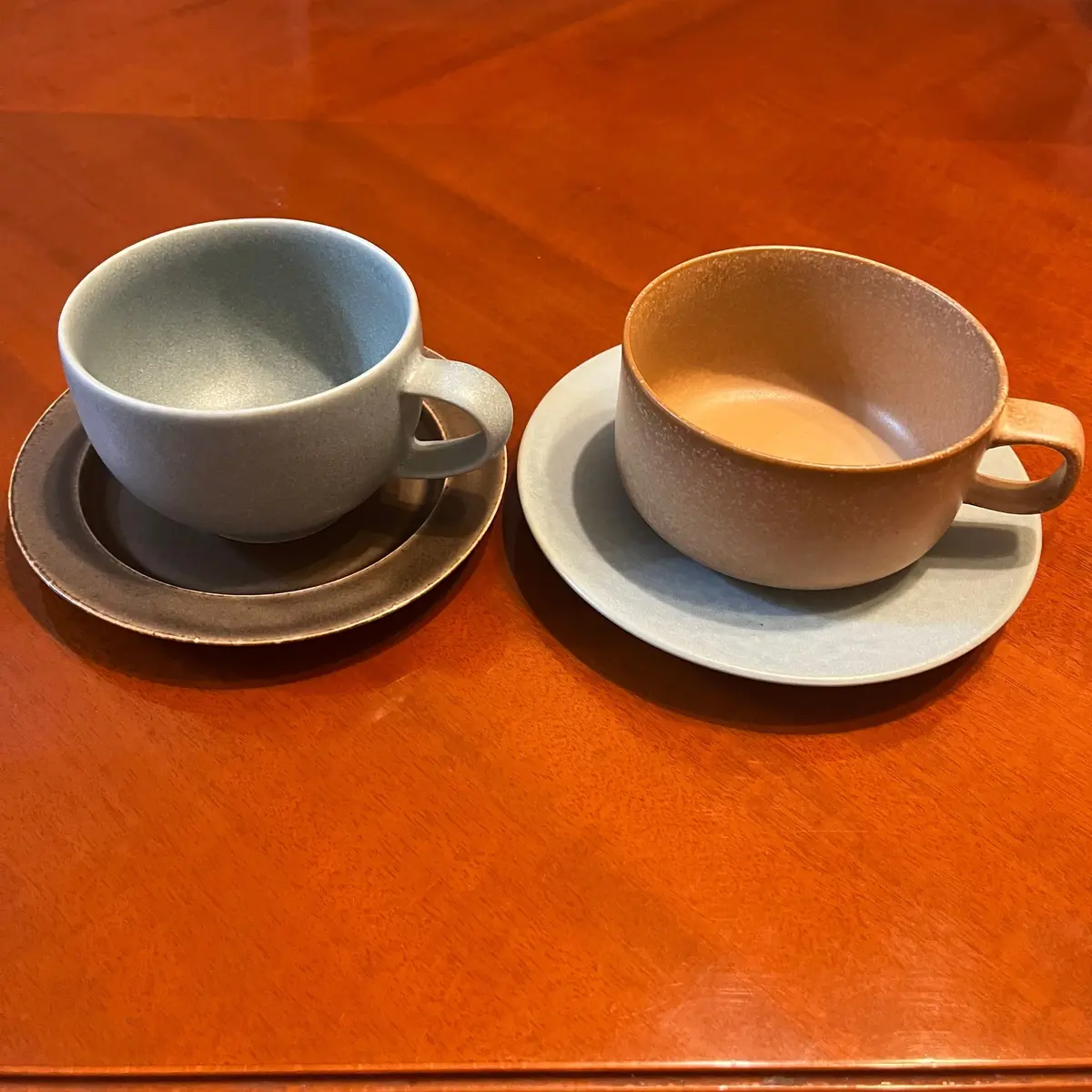 木製のテーブルに置かれた、2組の陶器のコーヒーカップとソーサー。左は青灰色、右はベージュと灰色のグラデーション。それぞれ異なる模様が施され、手作りの温かみを感じさせる。