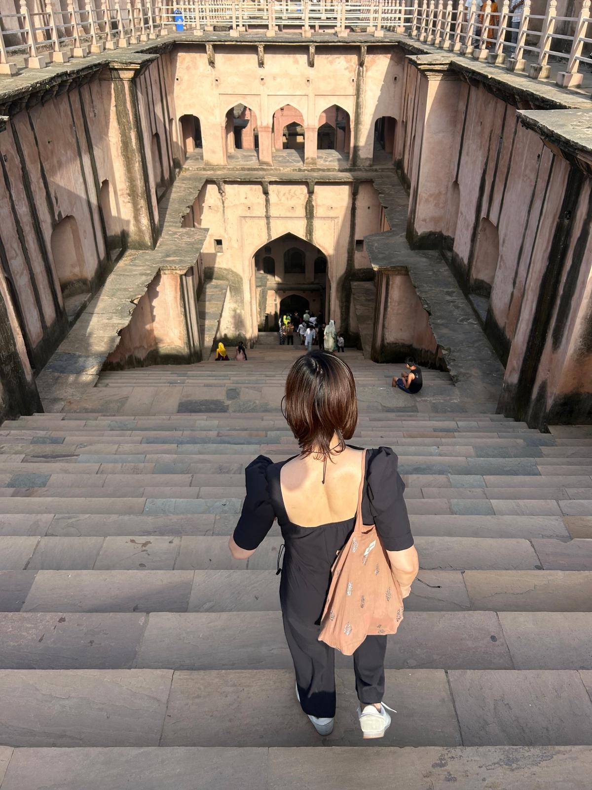 ピンク色の石造りの建造物と、その内部につながる長い石段。女性が背中を向け、石段を下りている。背景には、さらに奥へと続くアーチ型の通路が見える。