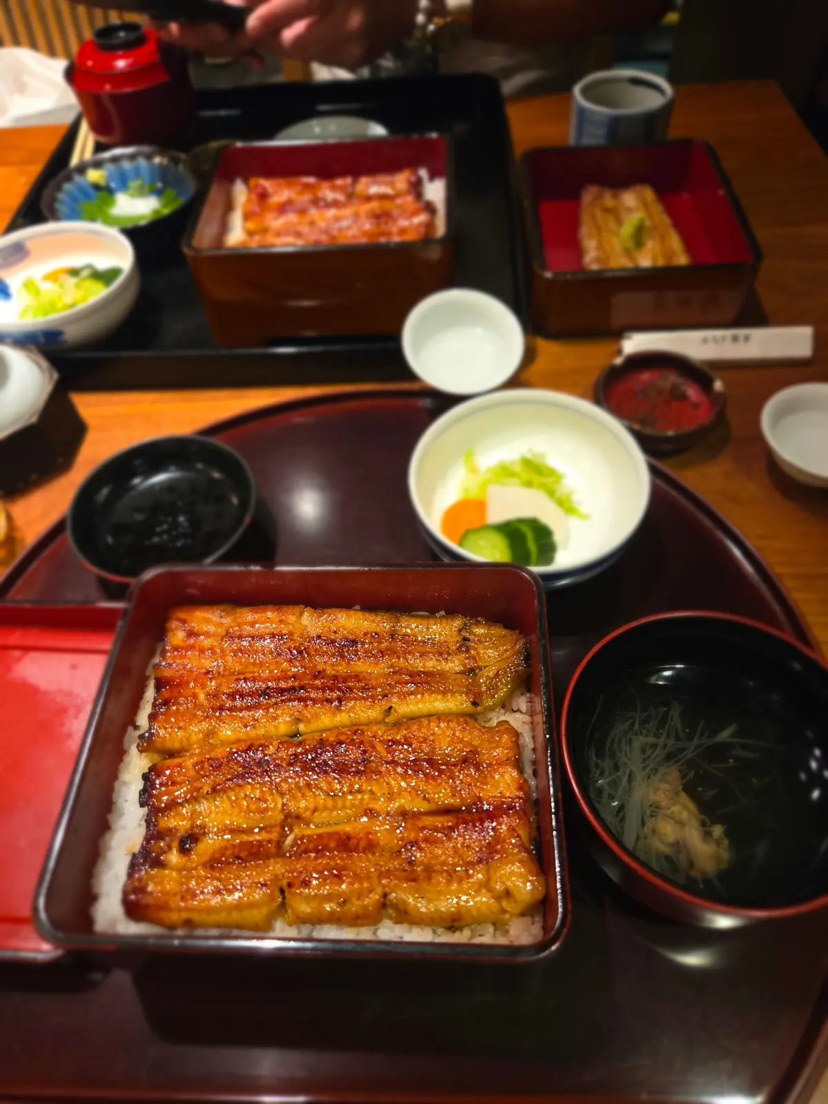 漆塗りの膳に、うなぎの蒲焼き丼、だし巻き卵、香の物、お吸い物などが盛り付けられている様子。うなぎは照り輝き、ご飯の上に並んでいる。