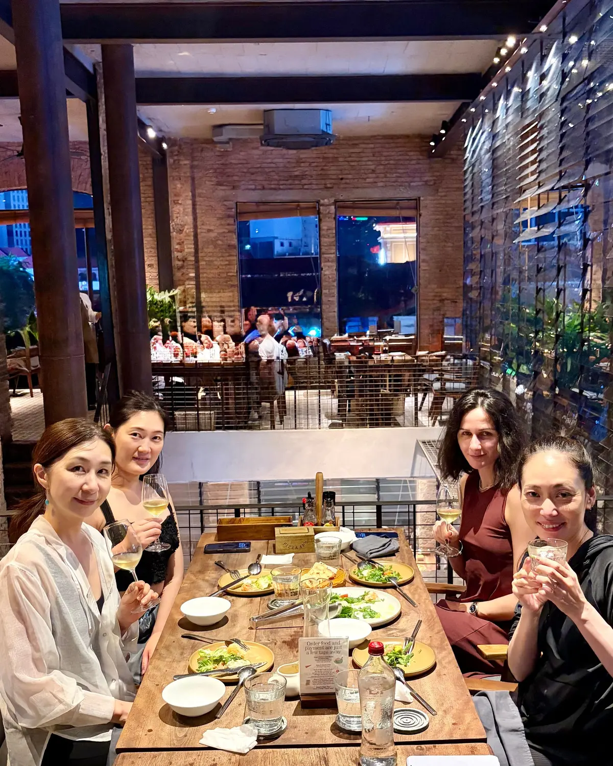 夜のレストランで4人の女性がテーブルに座り、食事と白ワインを楽しんでいる。テーブルには料理、グラス、ボトルが置かれ、背景には他の客人がいる。