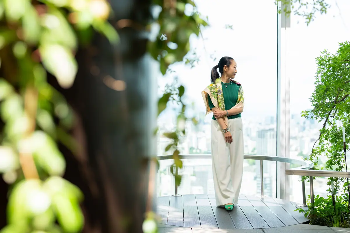 女性が屋上テラスに立ち、都市景観を眺めている。緑のトップスと白いパンツ、花柄のショールを着用。手は胸の前で組まれ、左手には時計が付いている。手前には緑の植物が配置され、背景には高層ビルが広がる。