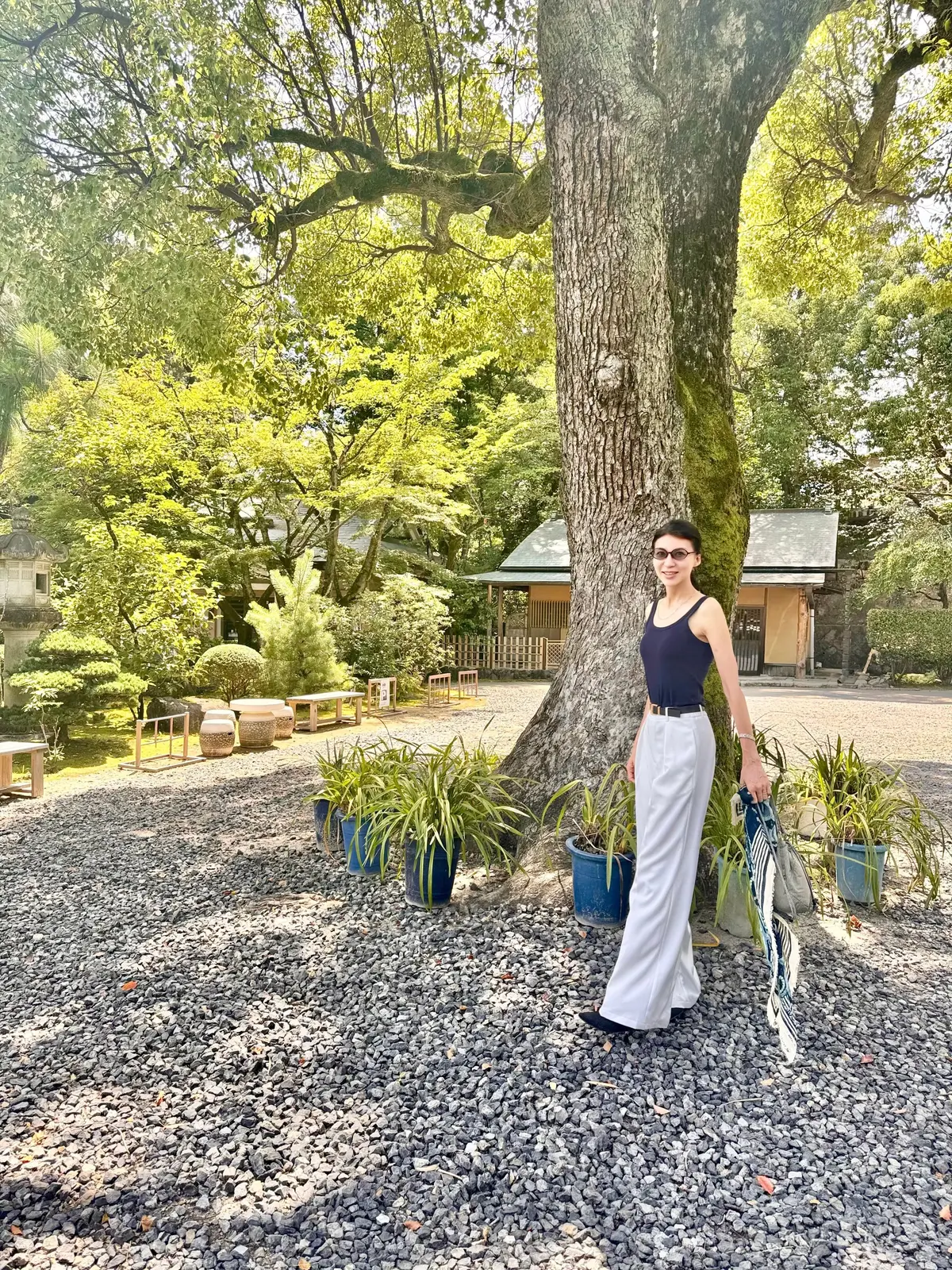 庭園に立つ女性。黒いタンクトップに白いパンツ、サングラスをかけ、右手にストールを持つ。背景には木々が生い茂り、伝統的な建物が見える。地面は小石敷き。