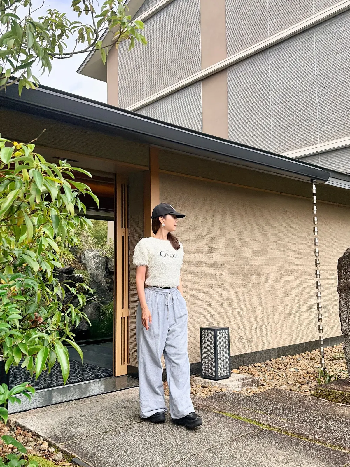 グレーのスウェット上下を着用した女性が、和風建築物の前に立っている。茶色のキャップを被り、右手を日差しから庇っている。背景には庭園が見える。