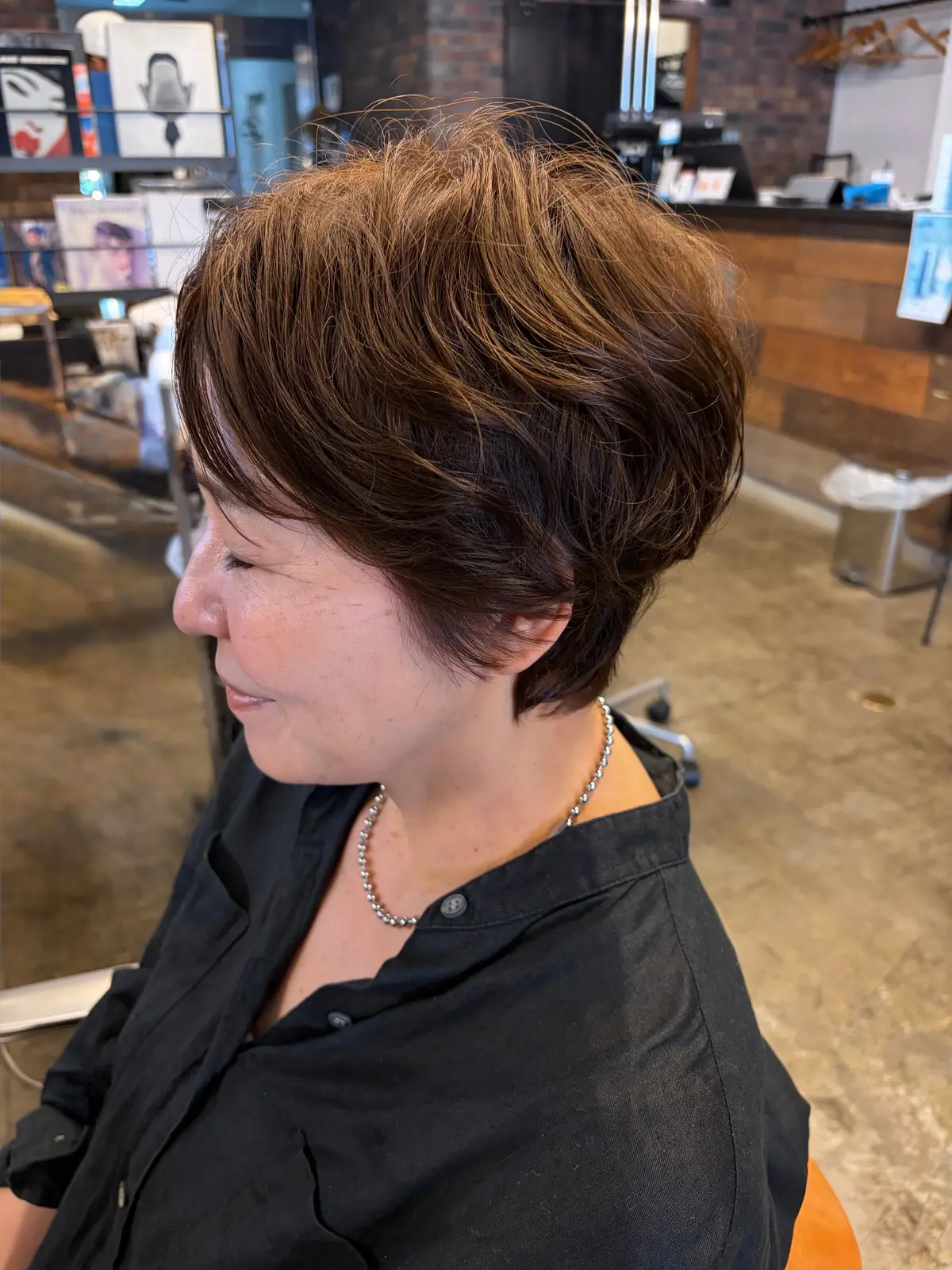 茶色のワックス加工されたショートヘアの女性。首には真珠のネックレスを着用。背景には理髪店の内装が見える。