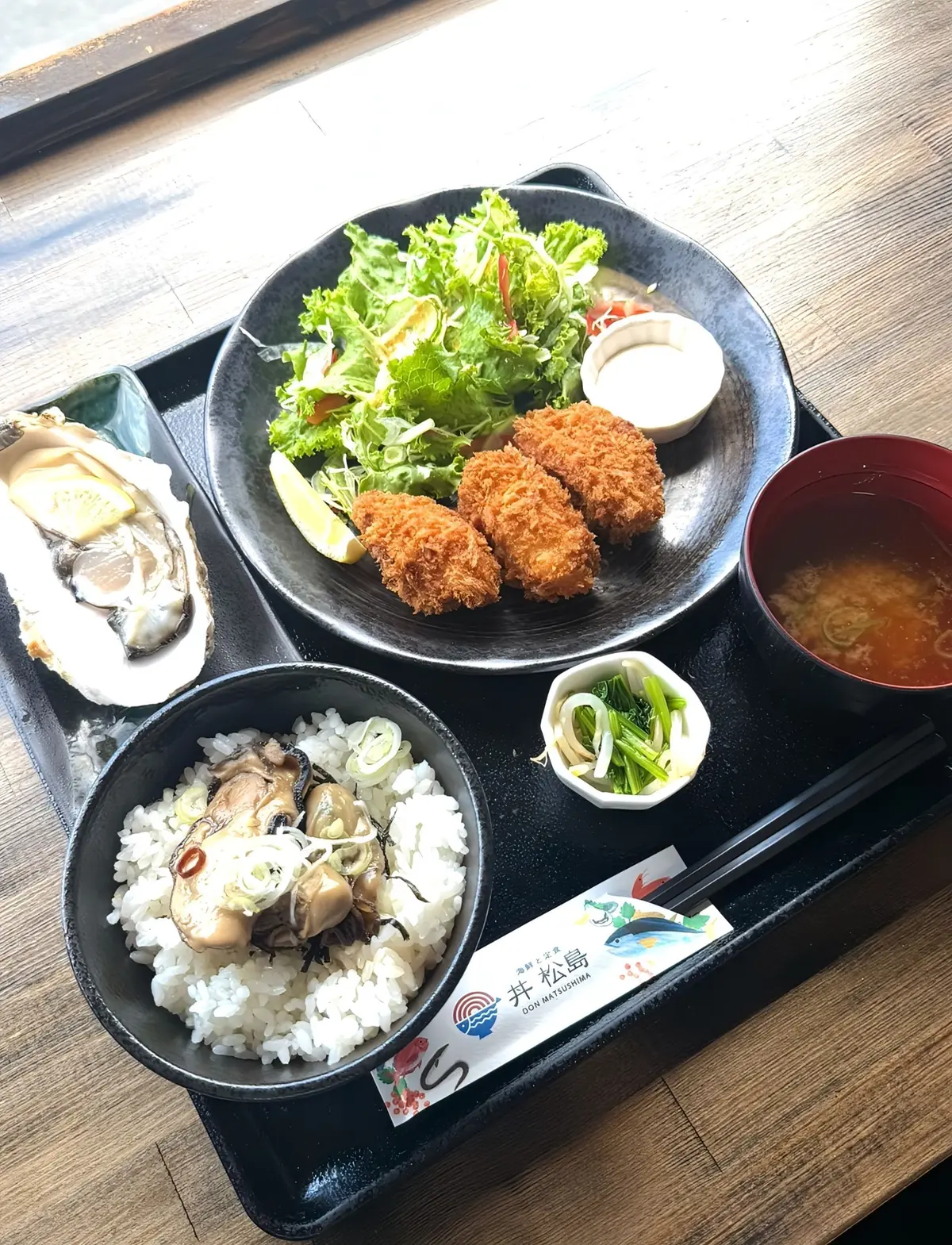 木製のテーブルに置かれた、日本の定食の盛り合わせ。中央にはサラダとコロッケ、左上にはカキ料理、左下には貝ご飯、右には赤色の汁物、右下には漬物と薬味が入った容器が置かれている。底には「丼 まつしま」のロゴが入った紙が敷かれている。