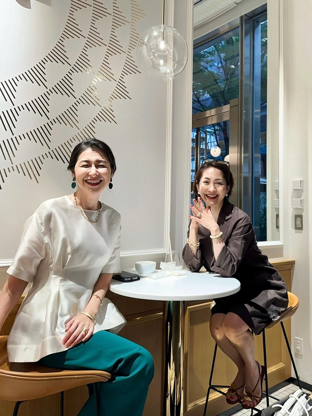 カフェのテーブルを挟んで座る2人の女性。左は白のトップスに緑のパンツ、右は茶色のシャツとスカート。笑顔でこちらを見ている。背景には幾何学模様の壁と窓がある。