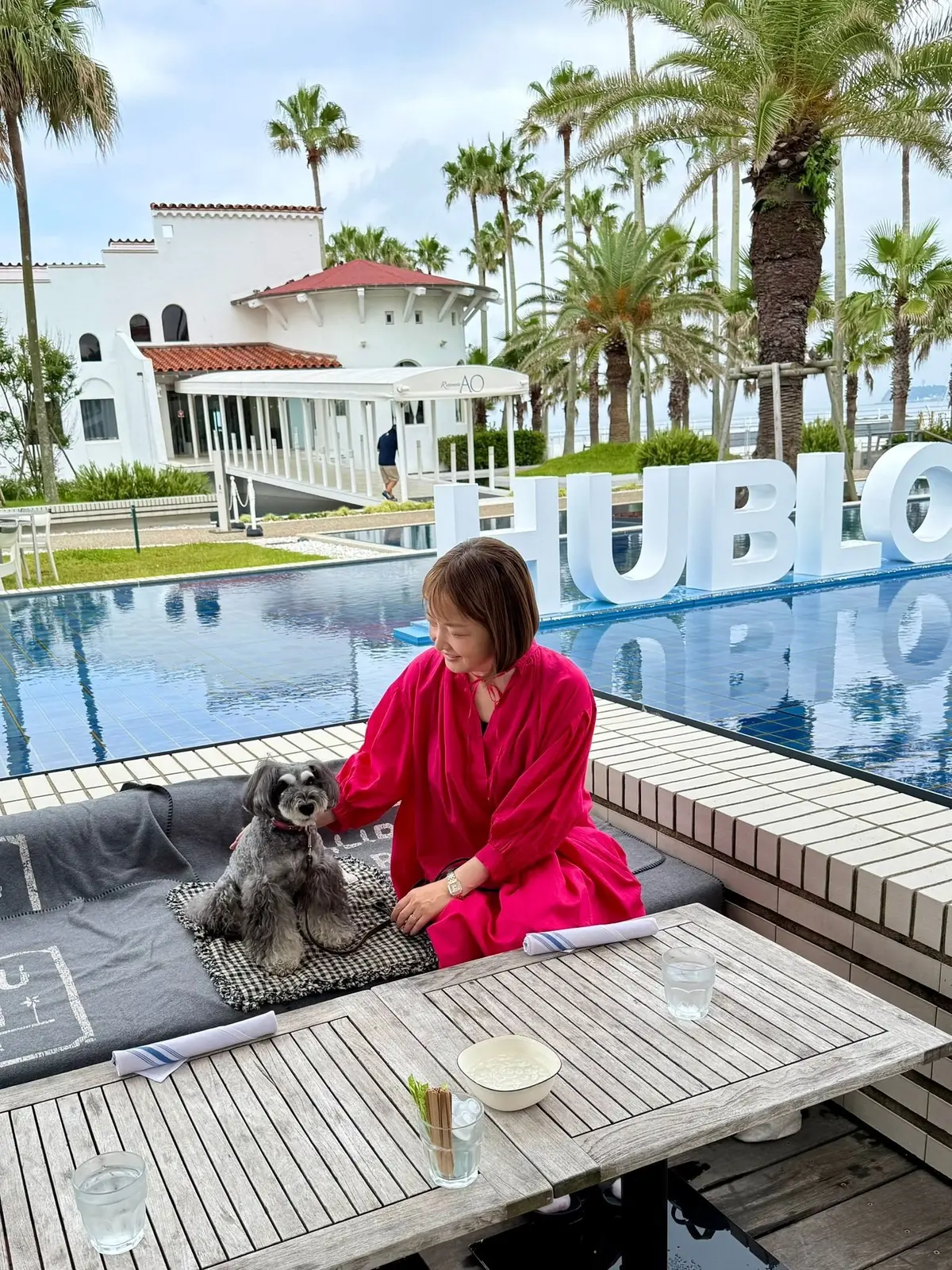 プールサイドのテーブルで、ピンクのシャツを着た女性が茶色の犬と触れ合っている。背景には白い建物とヤシの木、海が見える。