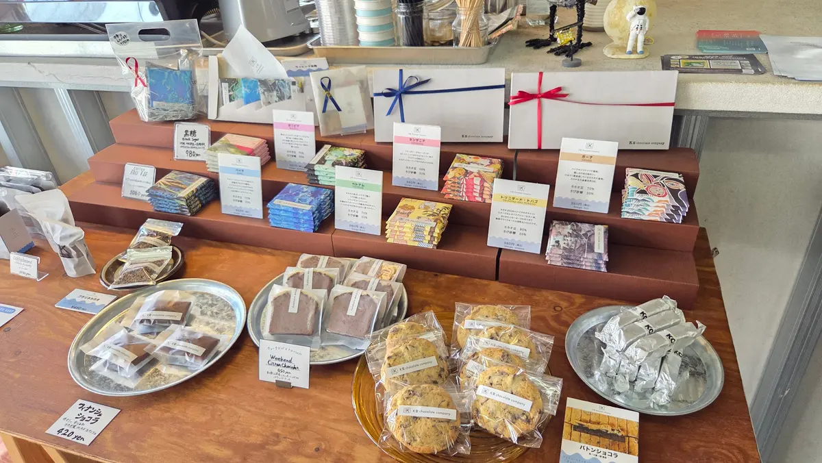 木製のテーブルに、チョコレートや焼き菓子、雑貨が並んでいる。棚には様々な種類のチョコレートが整然と並び、手前には金属製のトレイに焼き菓子が並べられている。背景には窓から光が差し込み、店内の一部が見える。