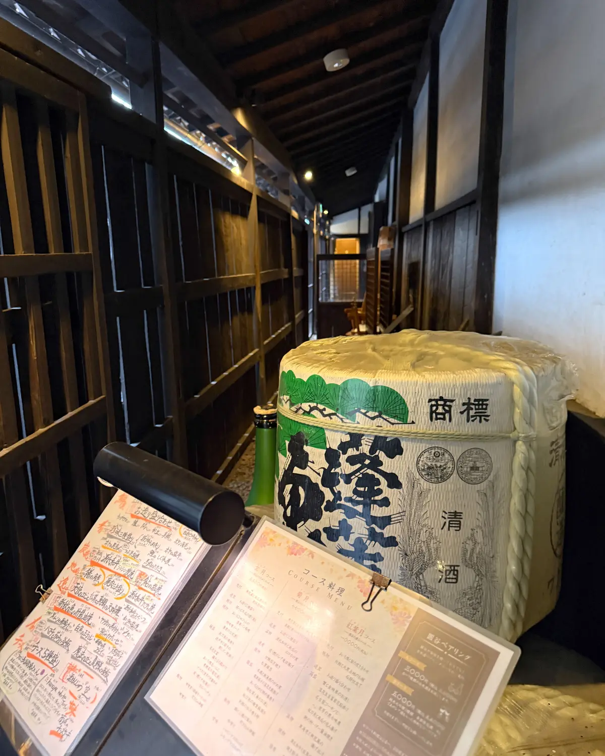 木造の狭い通路に置かれた、清酒の酒樽。樽には黒い文字で「商標 清酒」と書かれ、緑色の植物の模様が描かれている。樽の前にコースメニューが置かれている。