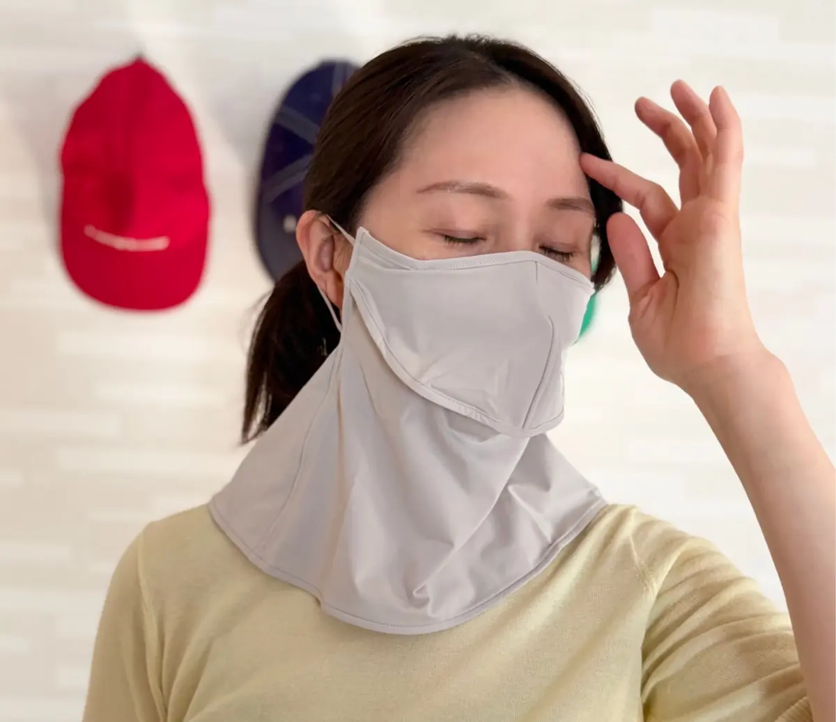 女性が白いマスクを着用し、右手で額を触れている。背景には赤と青の帽子が置かれている。