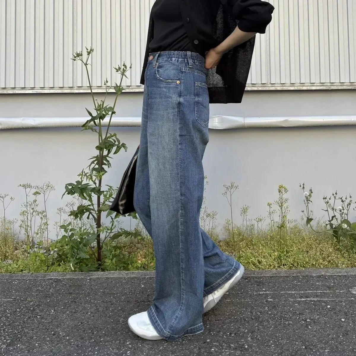 デニムパンツをはいた人物が、背景に白い壁と草むらがあり、黒の長袖シャツと黒のバッグを身につけています。