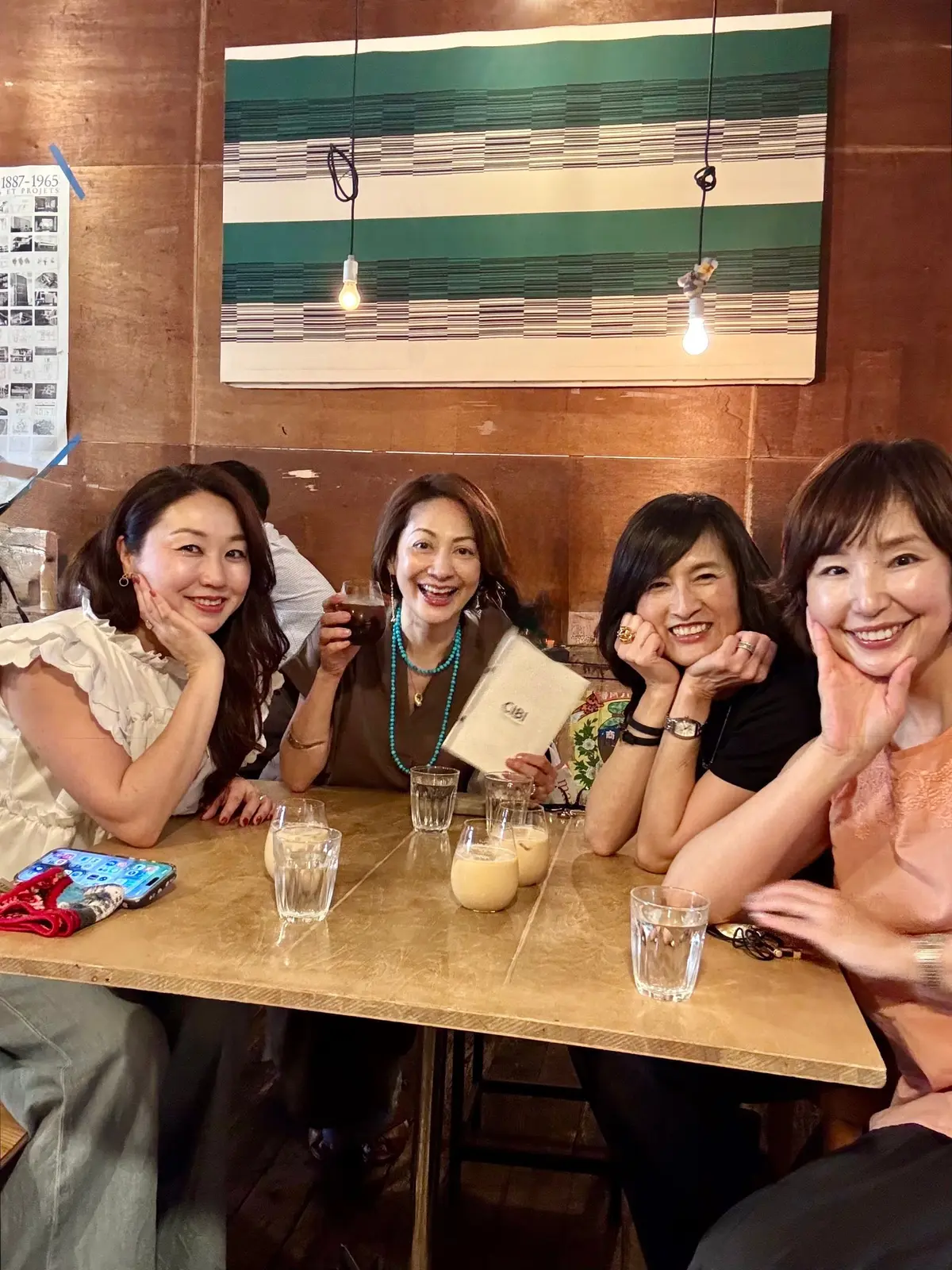 4人の女性が木製のテーブルに座り、笑顔で飲み物を楽しんでいる。中央の女性はメニューを手に持ち、左には携帯電話が置かれている。背景には緑と白の壁画と電球が吊るされている。