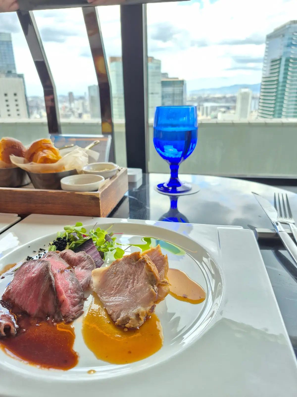 高層階の窓から都市の景色が見えるテーブルに、盛り付けられた肉料理、グラスに入った青色の飲み物、焼き菓子などが置かれている。