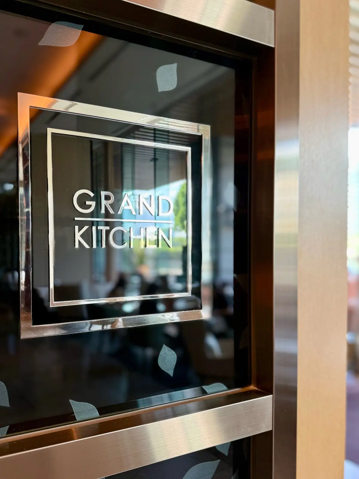 ブラス色のフレームに「GRAND KITCHEN」の文字が刻まれたサイン。背景はぼやけたレストランの様子。葉の形の装飾が左右に配置されている。