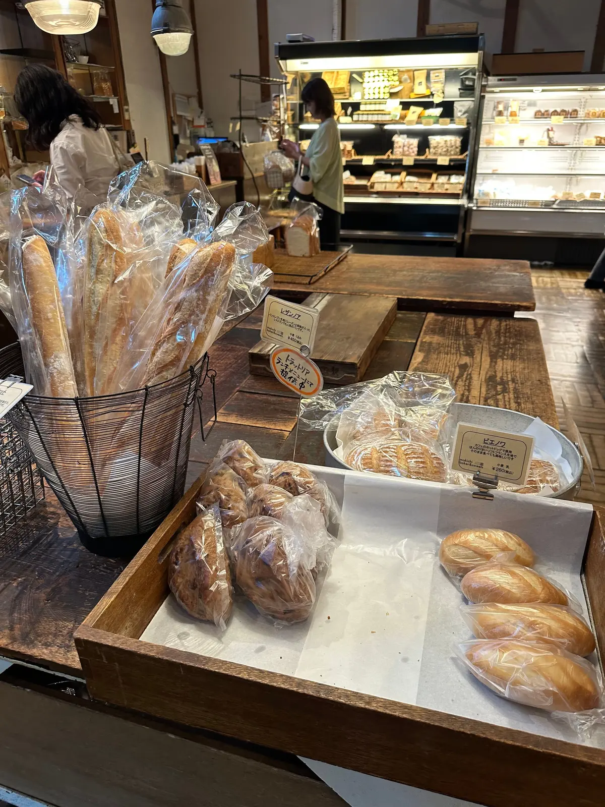 木製のテーブルに透明な袋に入ったパンが並び、店内にはパンの陳列棚や店員がいる