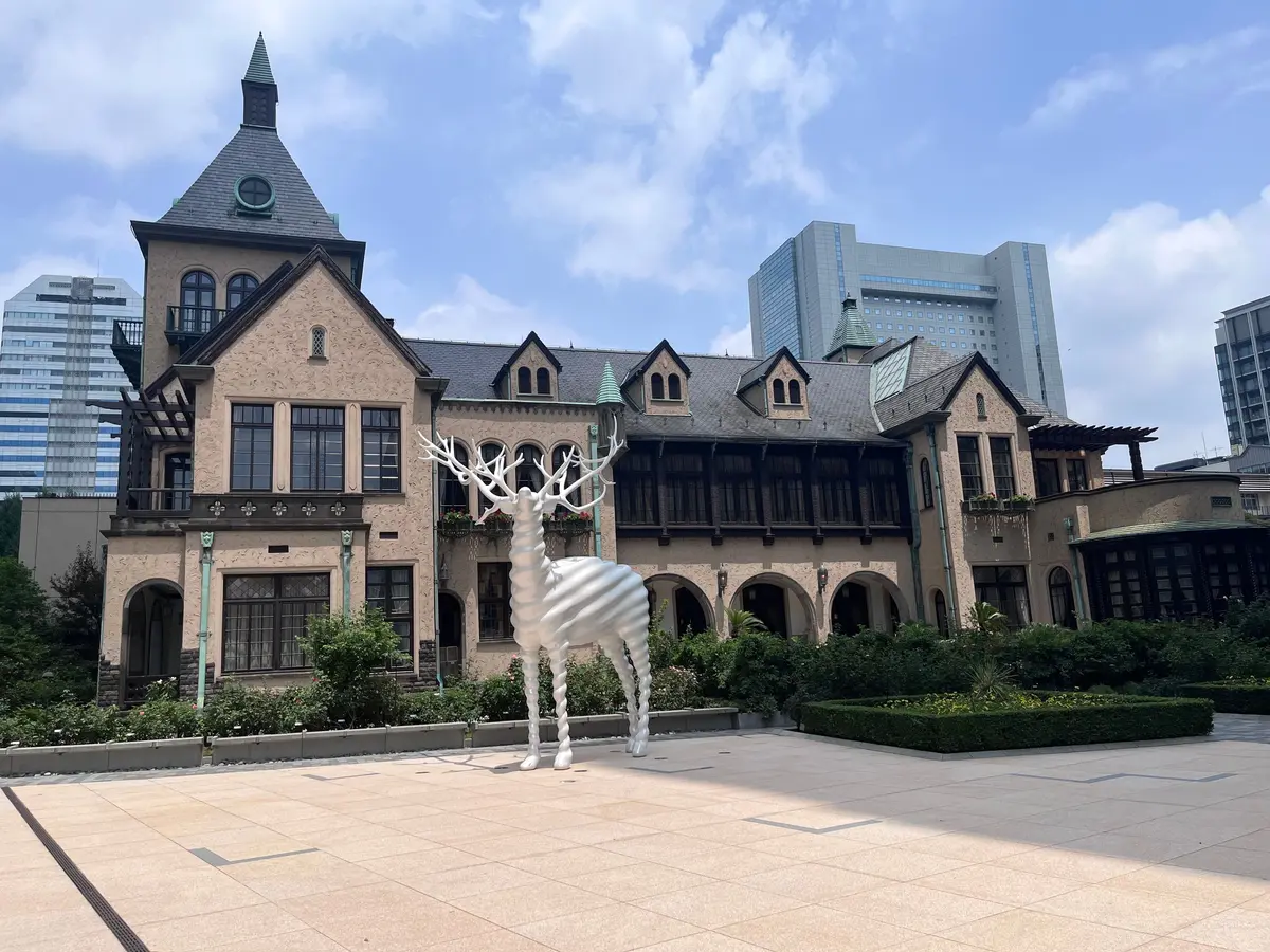 白い鹿のオブジェが置かれた、石畳の広場と歴史的な洋館の風景。背景には高層ビル群がそびえ立つ。
