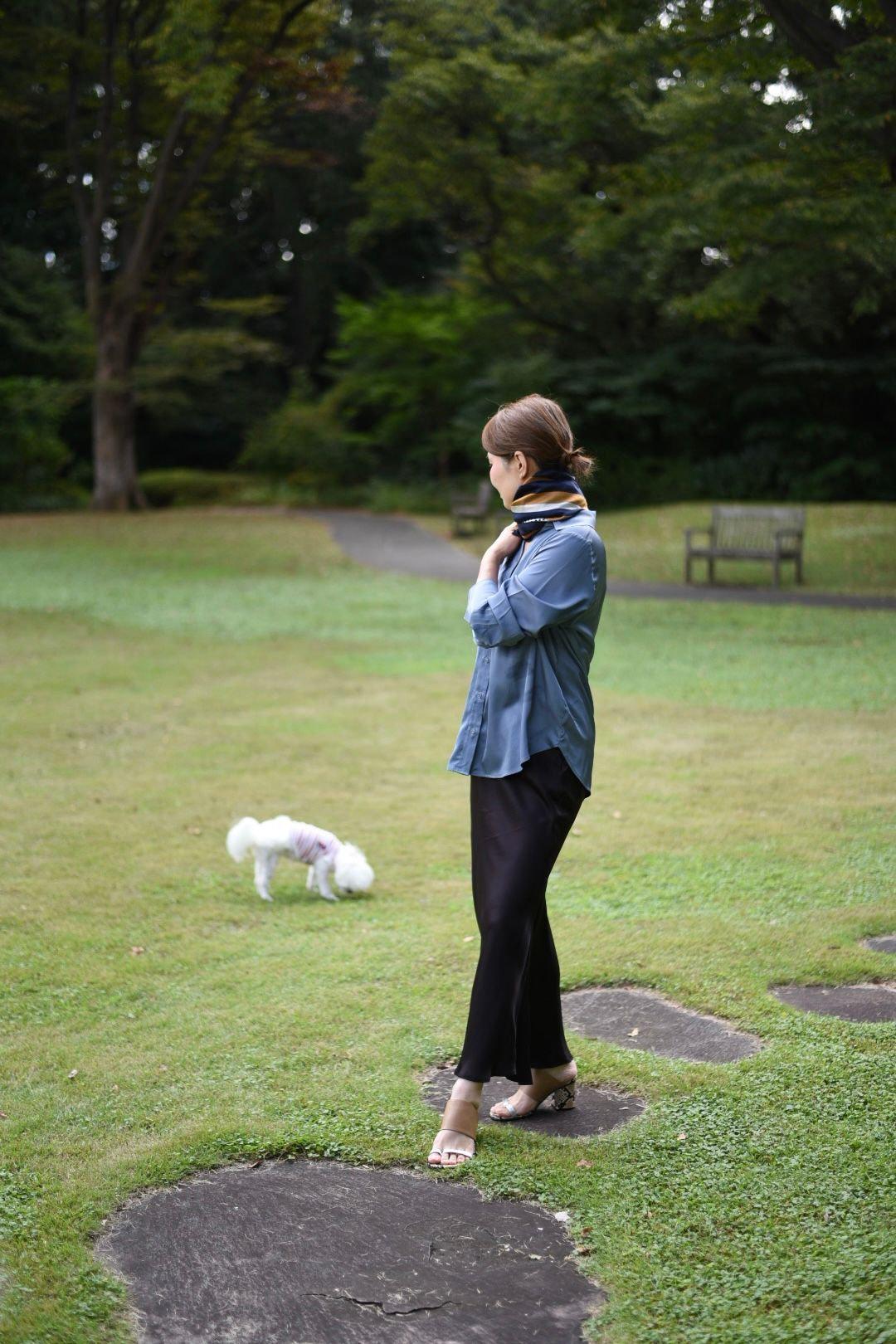 公園で、青いシャツに黒いパンツの女性が立っている。茶色の髪を後ろでまとめ、首にはスカーフを巻いている。白い犬が女性を見つめている。背景には木々とベンチが見える。
